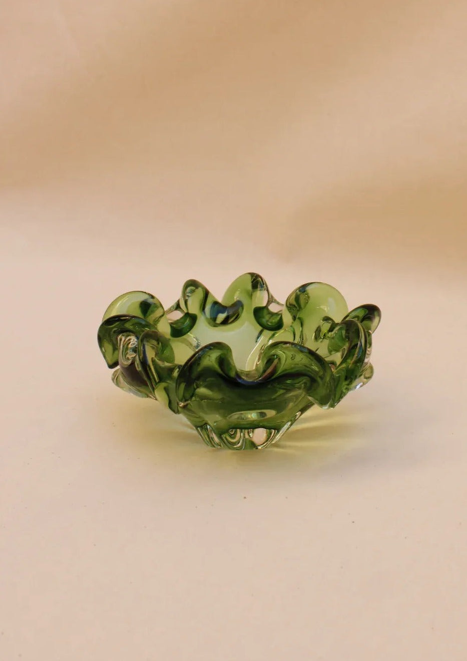 The Murano Glass Bowl from Boga Avante Shop is a small, handcrafted green glass piece with a wavy, ruffled rim. Its translucent design features light and dark green swirls, offering an artistic, fluid look ideal for luxury interiors or collectible furniture.