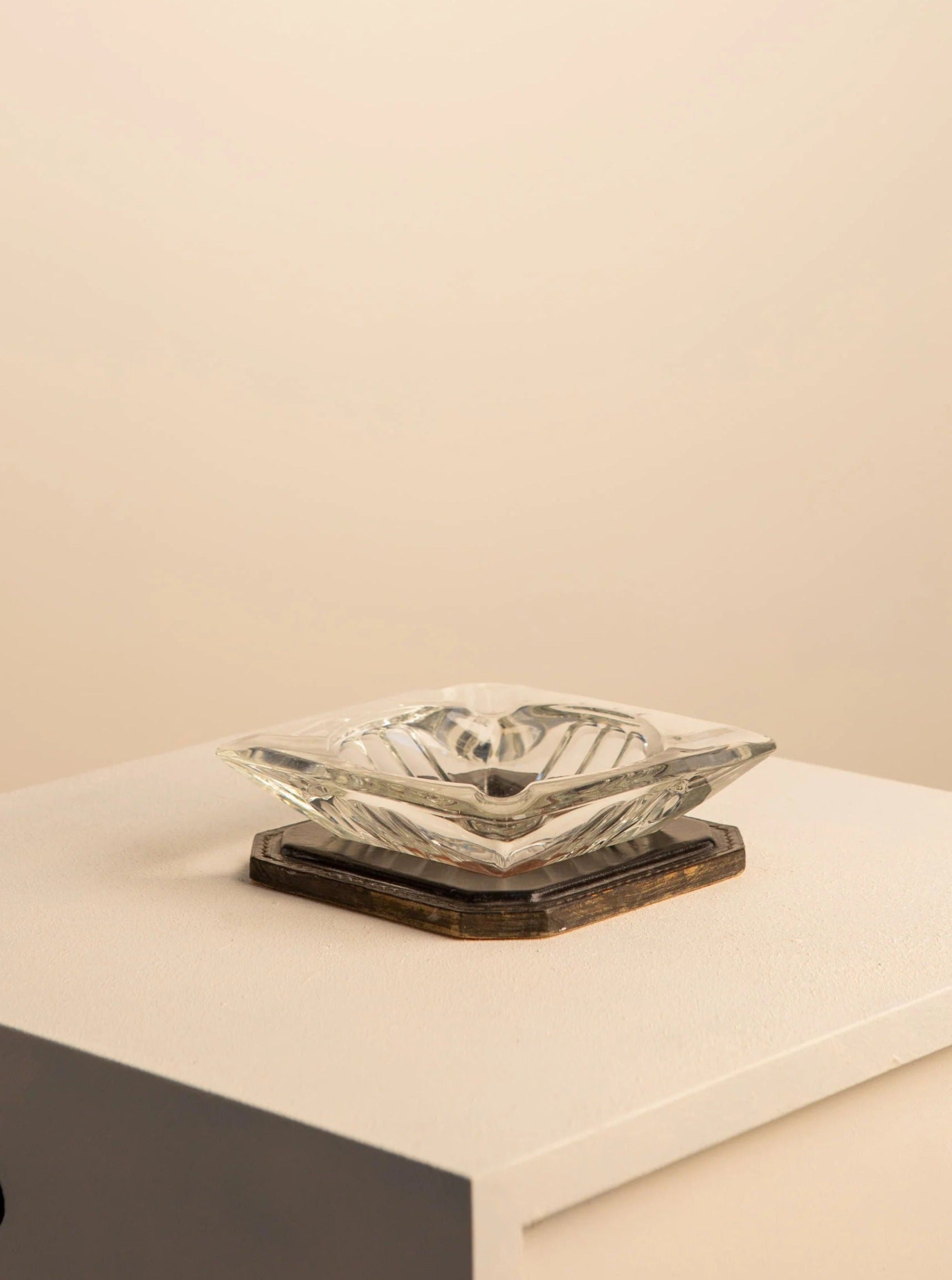 A sculptural Saint-Louis Crystal and Leather Ashtray by Treaptyque rests on a wooden coaster atop a cream table, accentuating contemporary design. Set against a neutral beige wall, the scene exudes an elegant and timeless ambiance.