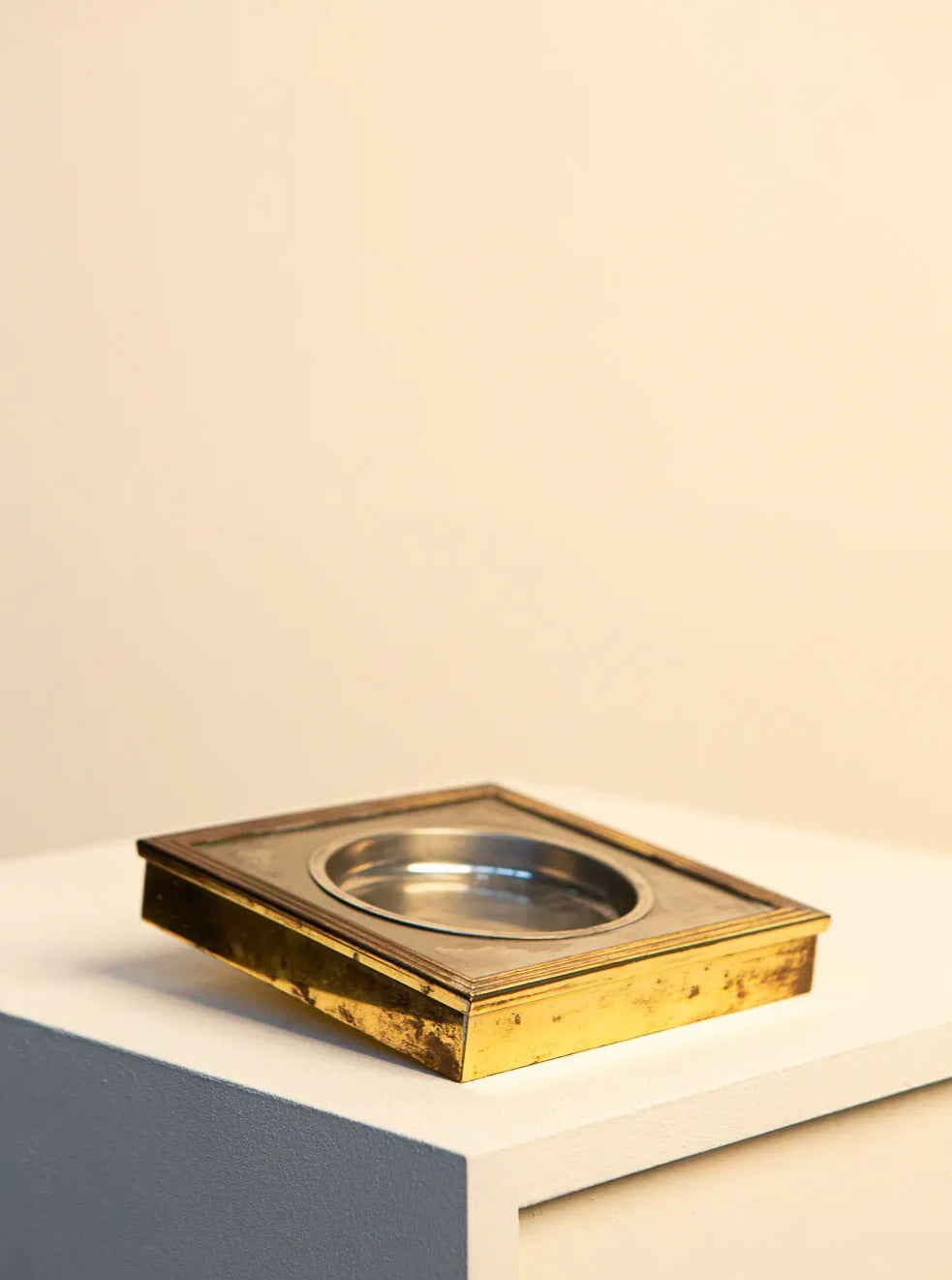 A 60's Silver and Gold-Plated Metal Square Tray with a clear glass center sits on a white pedestal against a beige background, casting a soft shadow by Treaptyque.
