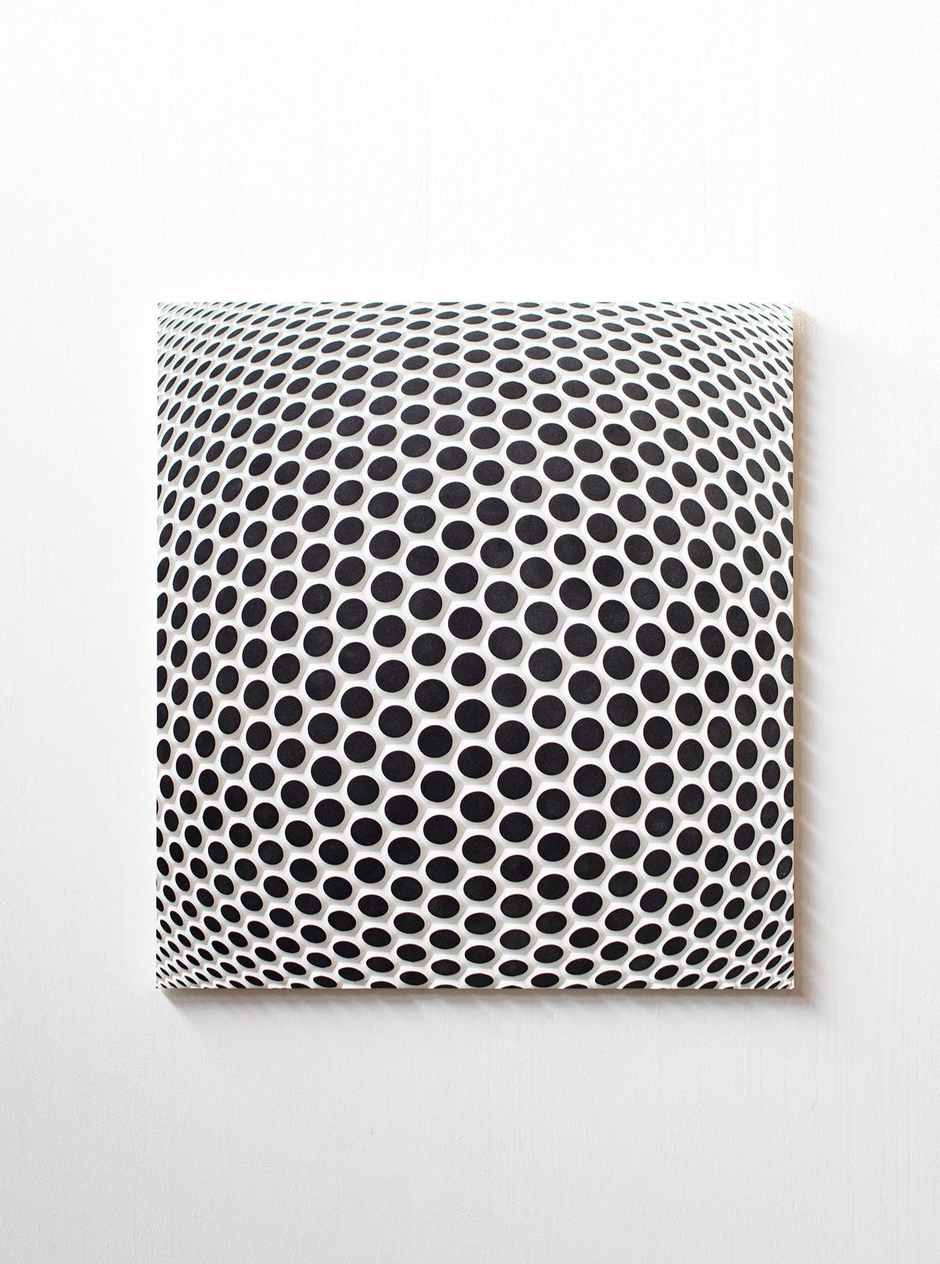 A Ommatidium art piece featuring a repeated pattern of black circles decreasing in size, giving an illusion of depth on a white square wooden wallpanel mounted on a white wall by Studio Arno Hoogland.