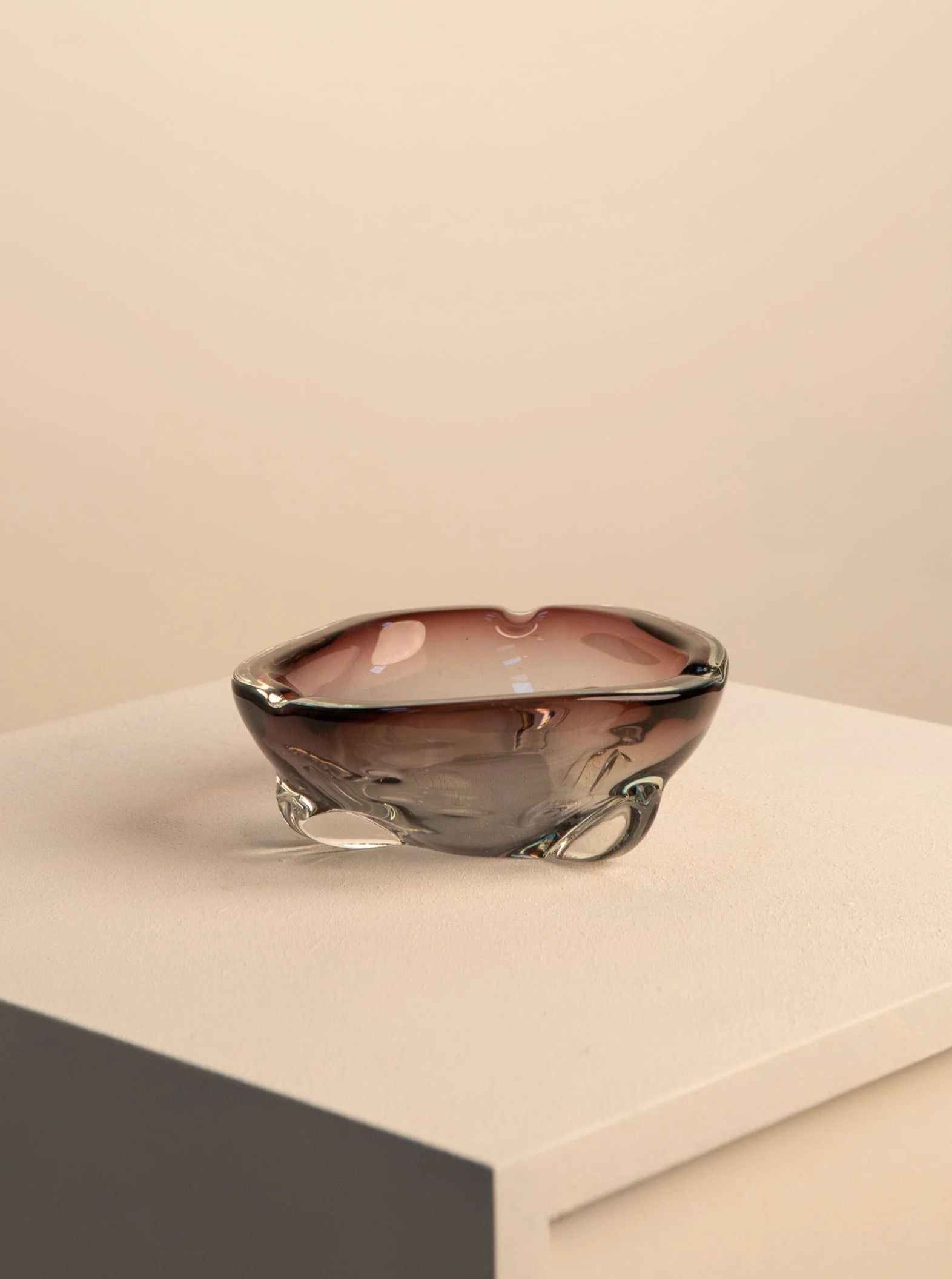 A small, translucent mauve Treaptyque glass bowl with a smooth, organic shape sitting on a light gray surface against a beige background.