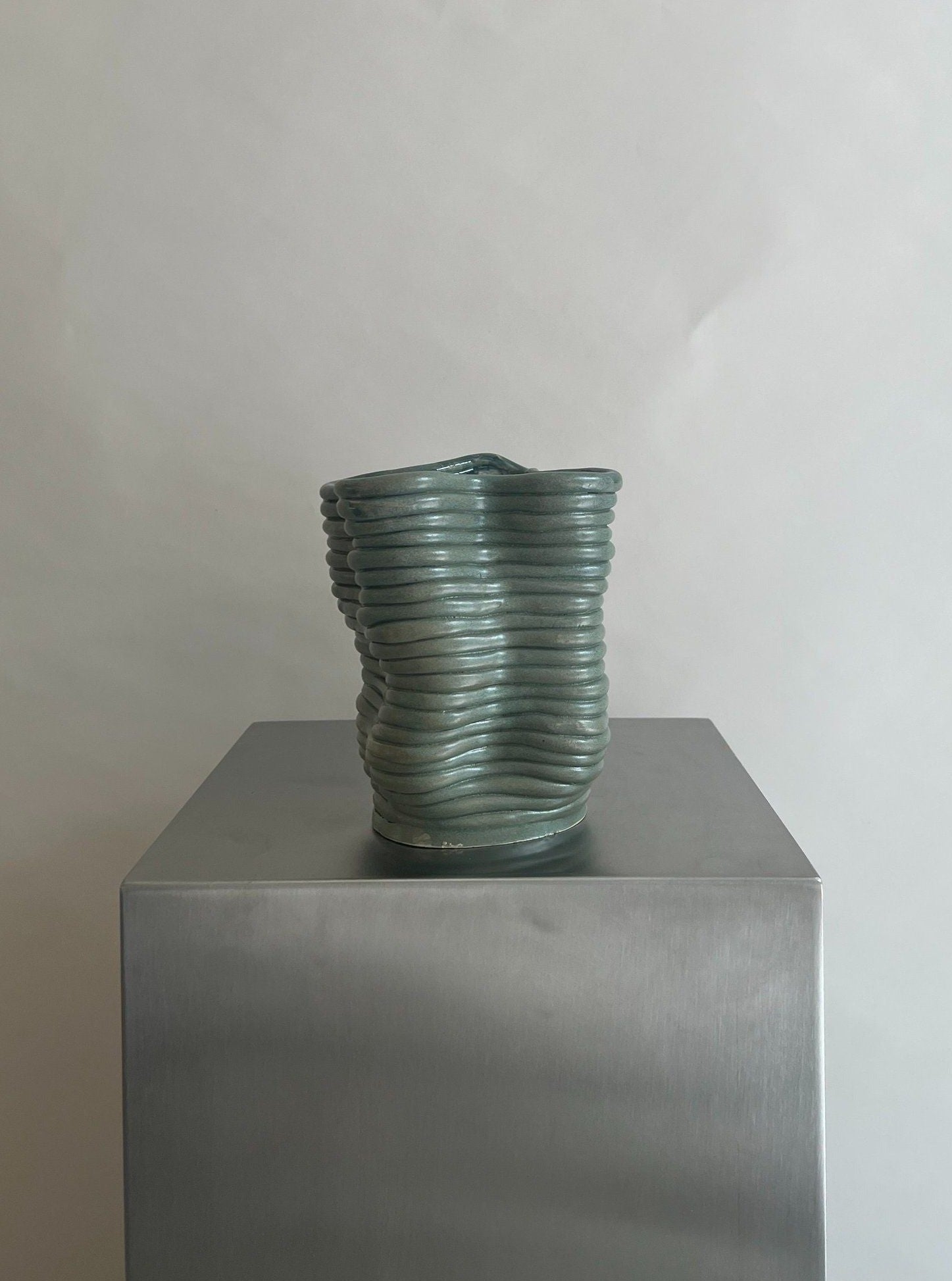 A handcrafted Wobbly Vase Green ceramic vase with a wavy, textured surface and a subtle green glaze sits atop a gray pedestal against a neutral background by 2222Studio.