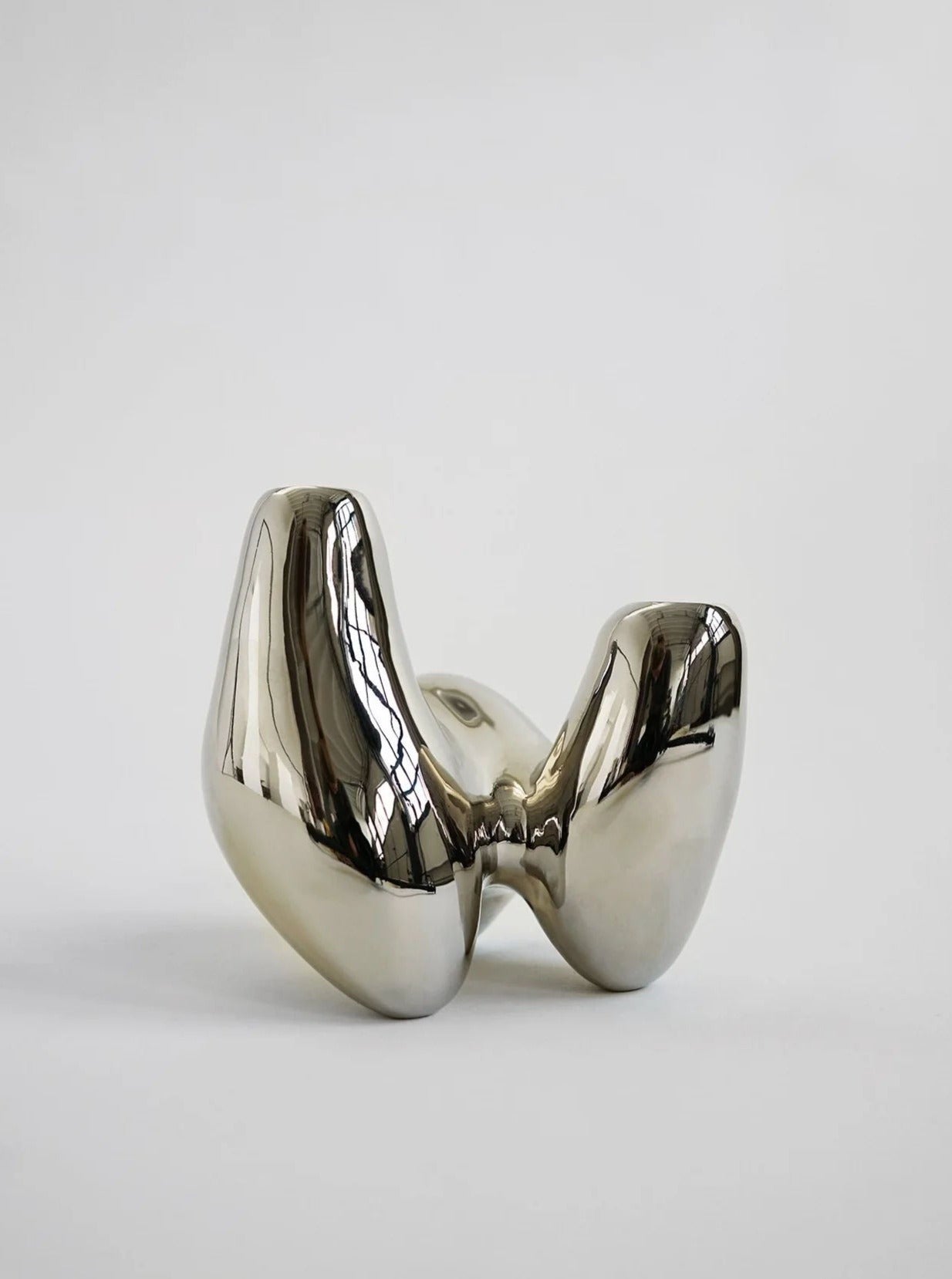 A polished, nickel-plated, abstract sculpture resembling a distorted letter "m" with a reflective surface, set against a plain white background by Hein Studio.
