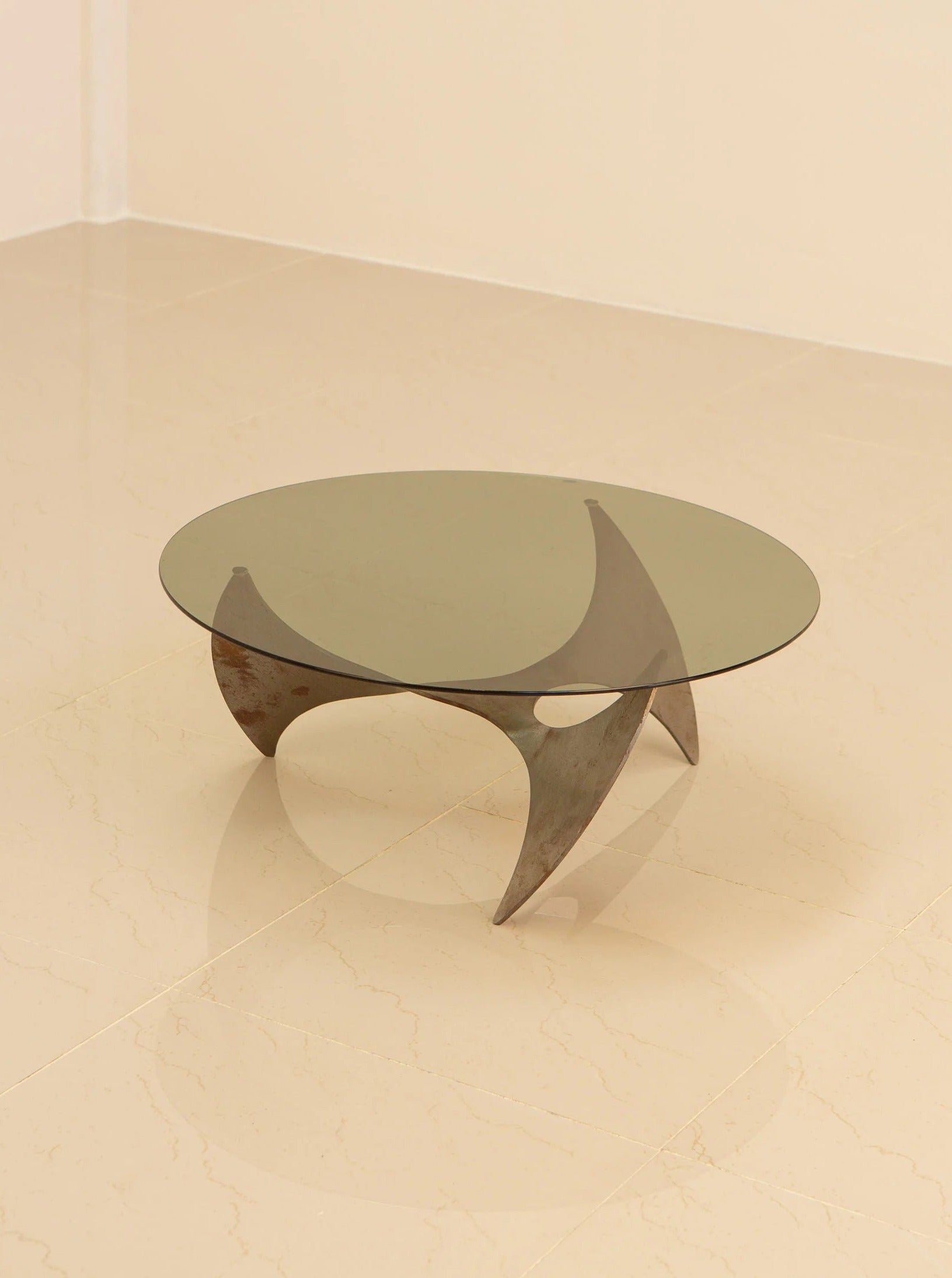 The Propeller Circular Coffee Table by Knut Hesterberg, crafted by Treaptyque, elegantly adorns the polished beige tile floor of a minimalist room. This round glass table boasts a unique and curved wooden base with an intersecting support structure that exudes a sleek and intricate design reminiscent of Hesterberg's iconic style.