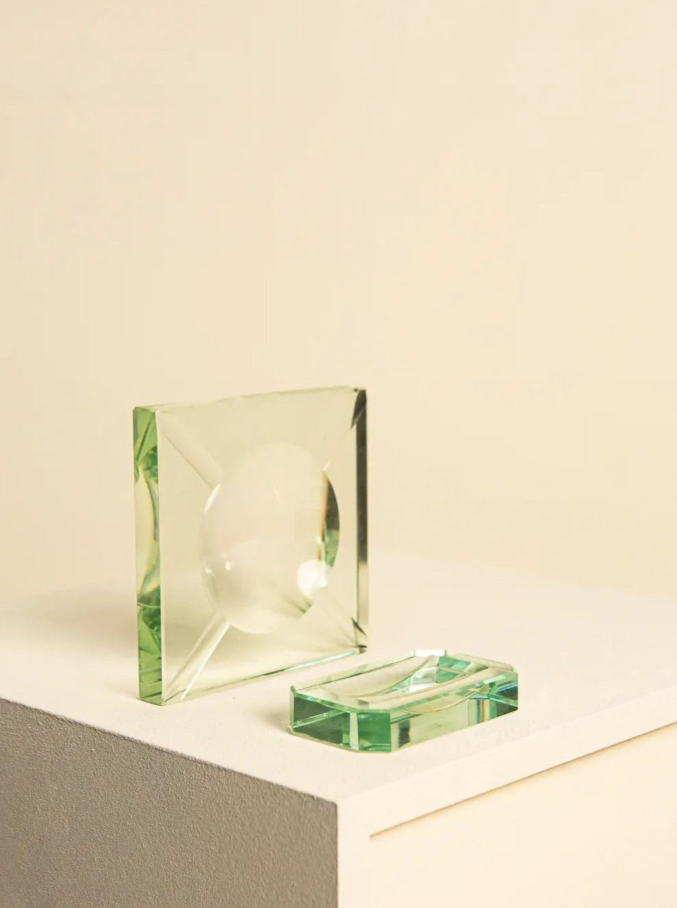 A square glass object with beveled edges and a spherical indent rests on a white surface. Next to it lies a rectangular glass piece with rounded edges, placed on the same surface. Both pieces are part of the vintage 60's Glass Desk Set by Fontana Arte, offered by Treaptyque, crafted from clear, light green-tinted glass that evokes Italian design aesthetics.