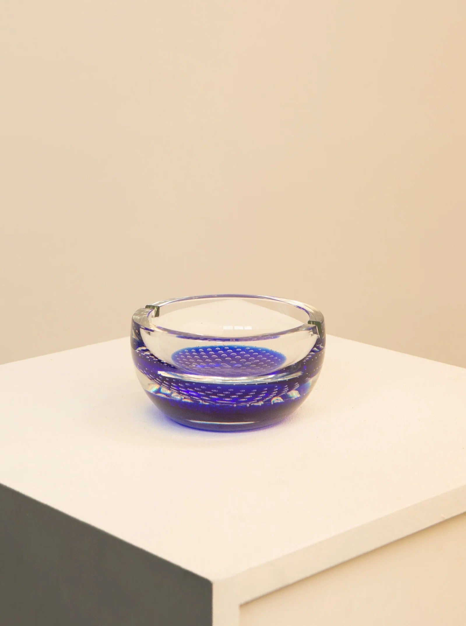 A clear Treaptyque 70's Blue Murano Glass Vide Poche with a blue dotted pattern on a light beige surface, highlighted by subtle lighting, creating a calm and minimalist aesthetic.