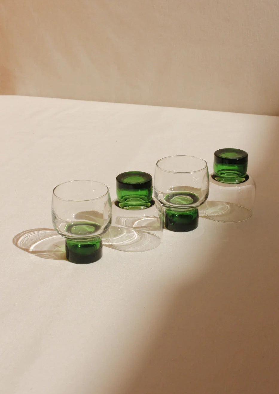 Five transparent art deco French Mid-Century Glasses with green bottoms and handles on a cream background, elegantly arranged in a staggered format. Two glasses are flipped upside down. (Brand Name: Boga Avante Shop)