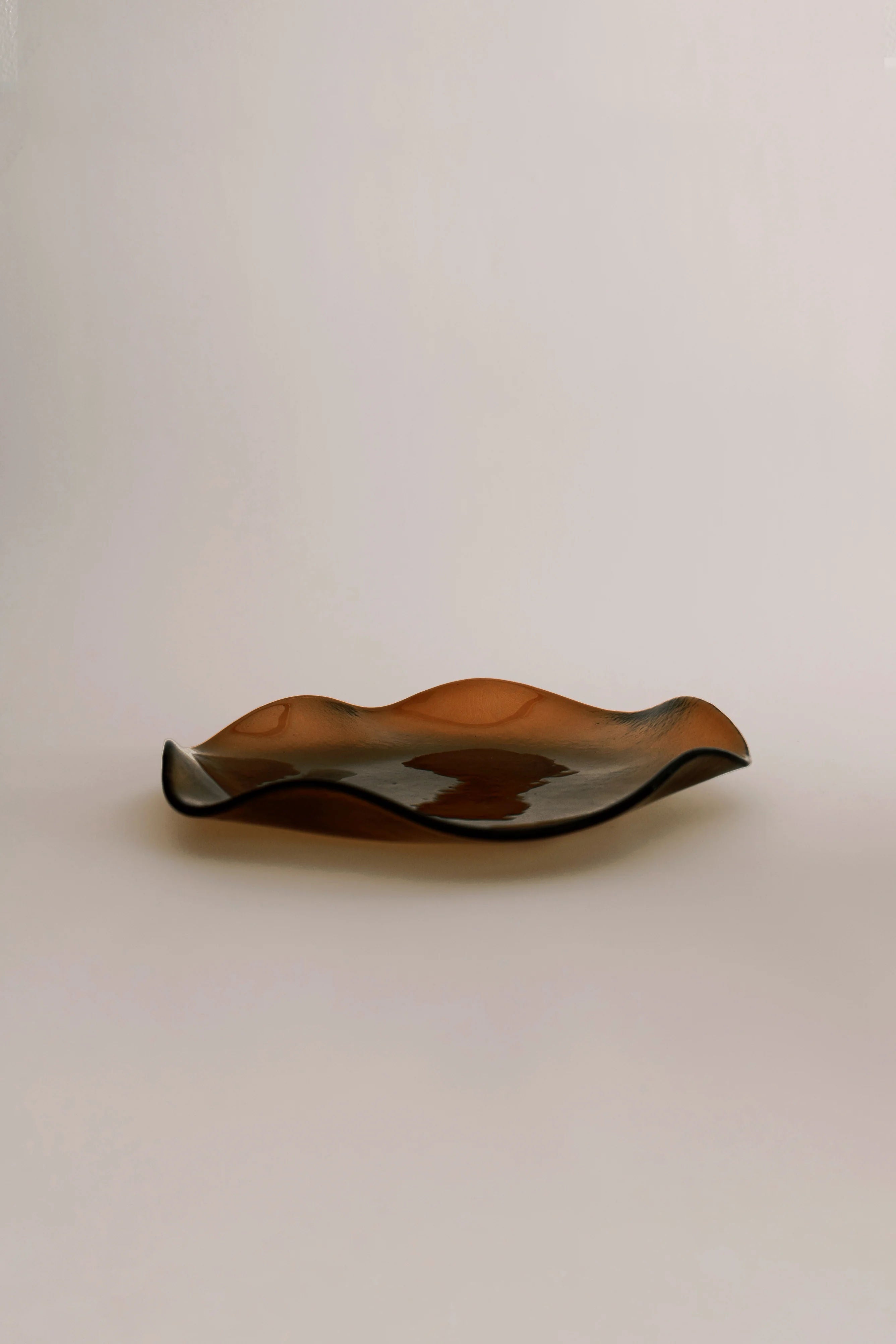 A sleek, undulating glass Petal Plate - Large with a glossy finish, set against a neutral background, displaying earthy brown tones with subtle reflections from its artisanal production process by Sophie Lou Jacobsen.