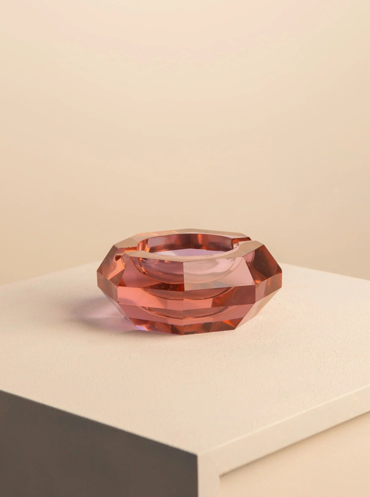 A large, faceted pink Treaptyque Murano glass ashtray rests on a gray surface against a soft beige background, reflecting light through its translucent surface.