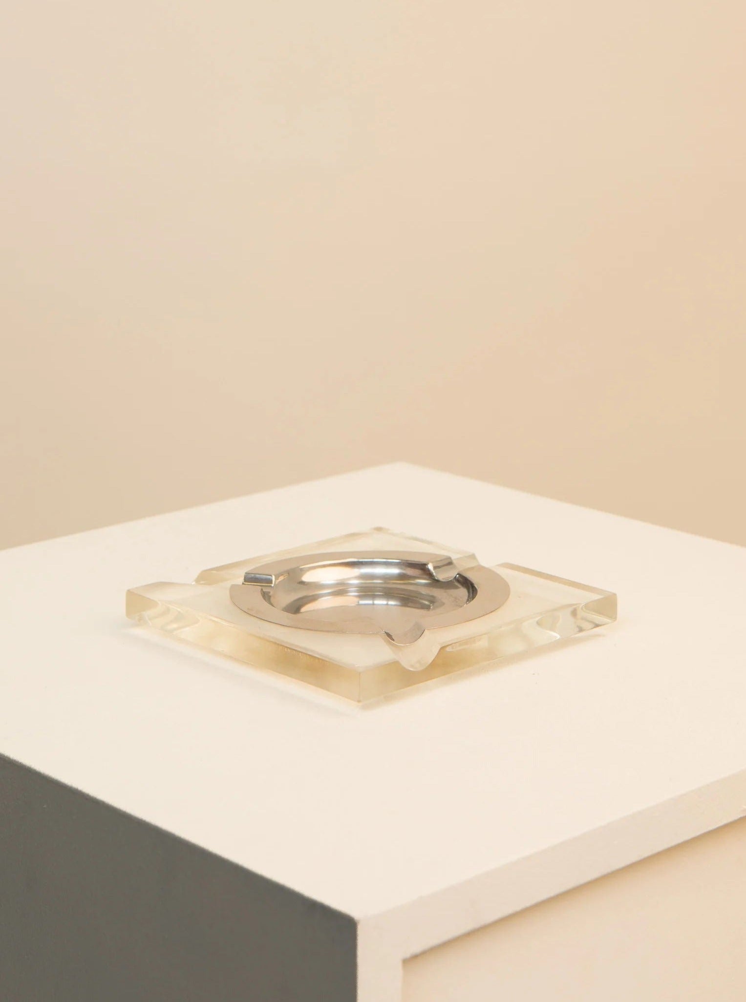 A minimalist photograph featuring a Treaptyque Plexiglass and Metal 70's Ashtray with a geometric design, placed on a white surface against a soft beige background.