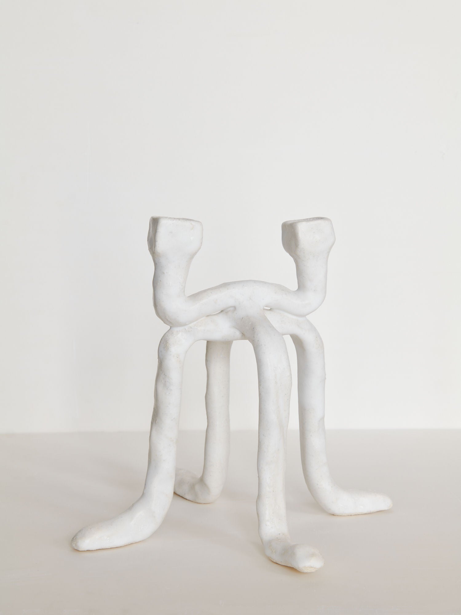 A white chamotte stoneware sculpture resembling a chair with elongated, irregular legs and rounded armrests, displayed against a plain background by Candleholder La Chose 2 from Claire Pain.