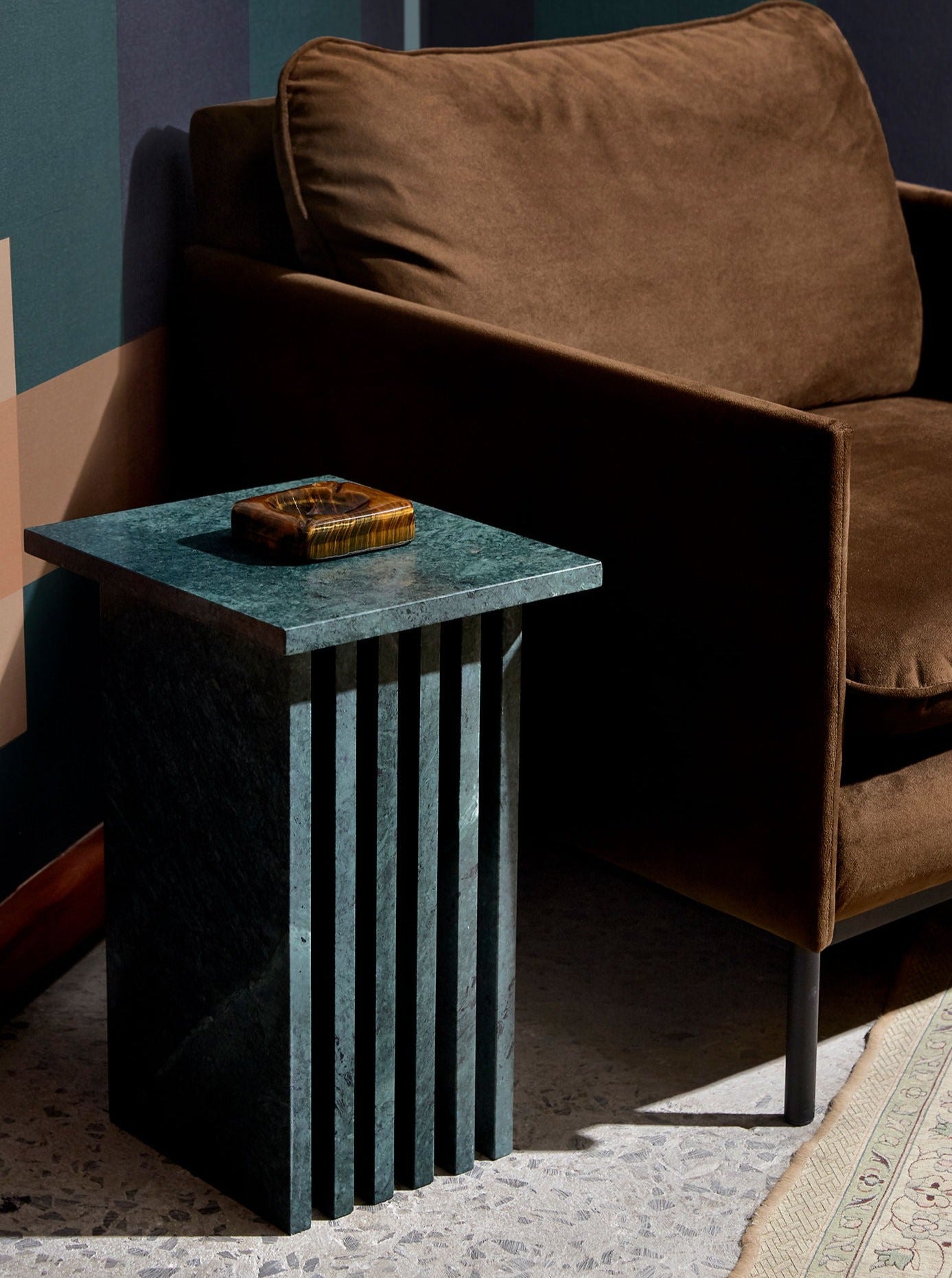 A modern living room with a plush brown sofa and a stylish Lemon Vondel Side Table in Verde Guatemala topped with a green marble slab. A small decorative item is on the table, bathing in natural light.