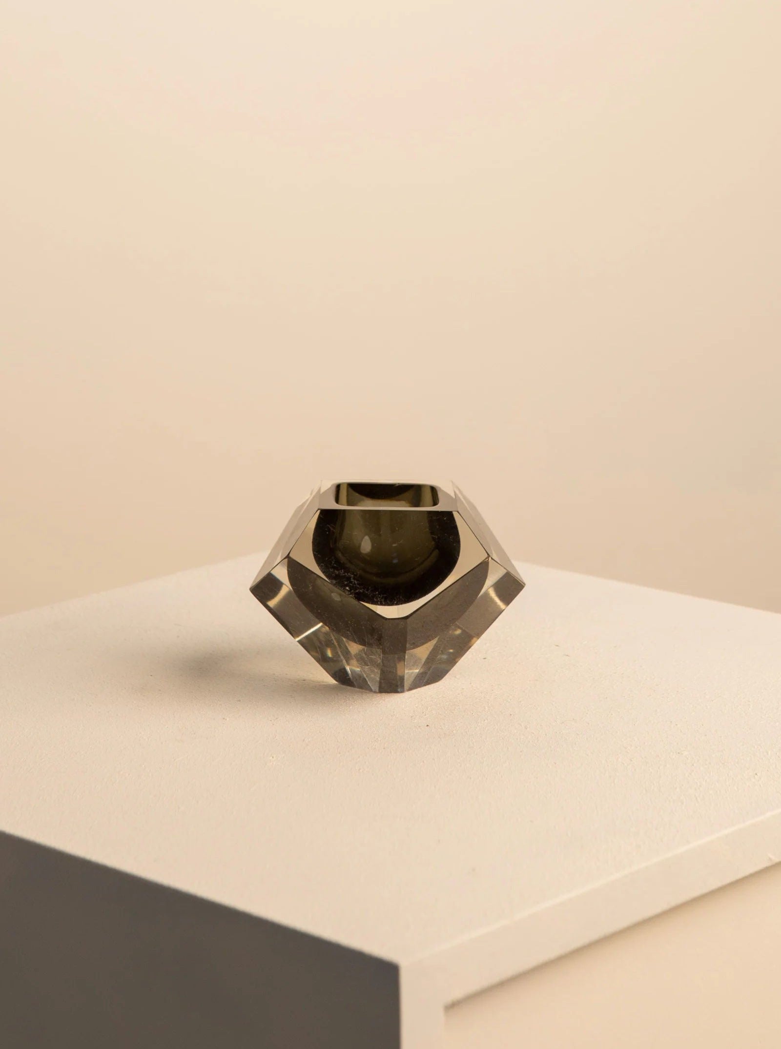 A black "Diamant" ashtray in Murano glass by Flavio Poli for Seguso from the 60s, offered by Treaptyque, sits on a white pedestal. Its small geometric design features sharp, angled facets and a transparent base with a slight grey tint, casting subtle shadows on the pedestal and background to create a minimalist aesthetic.