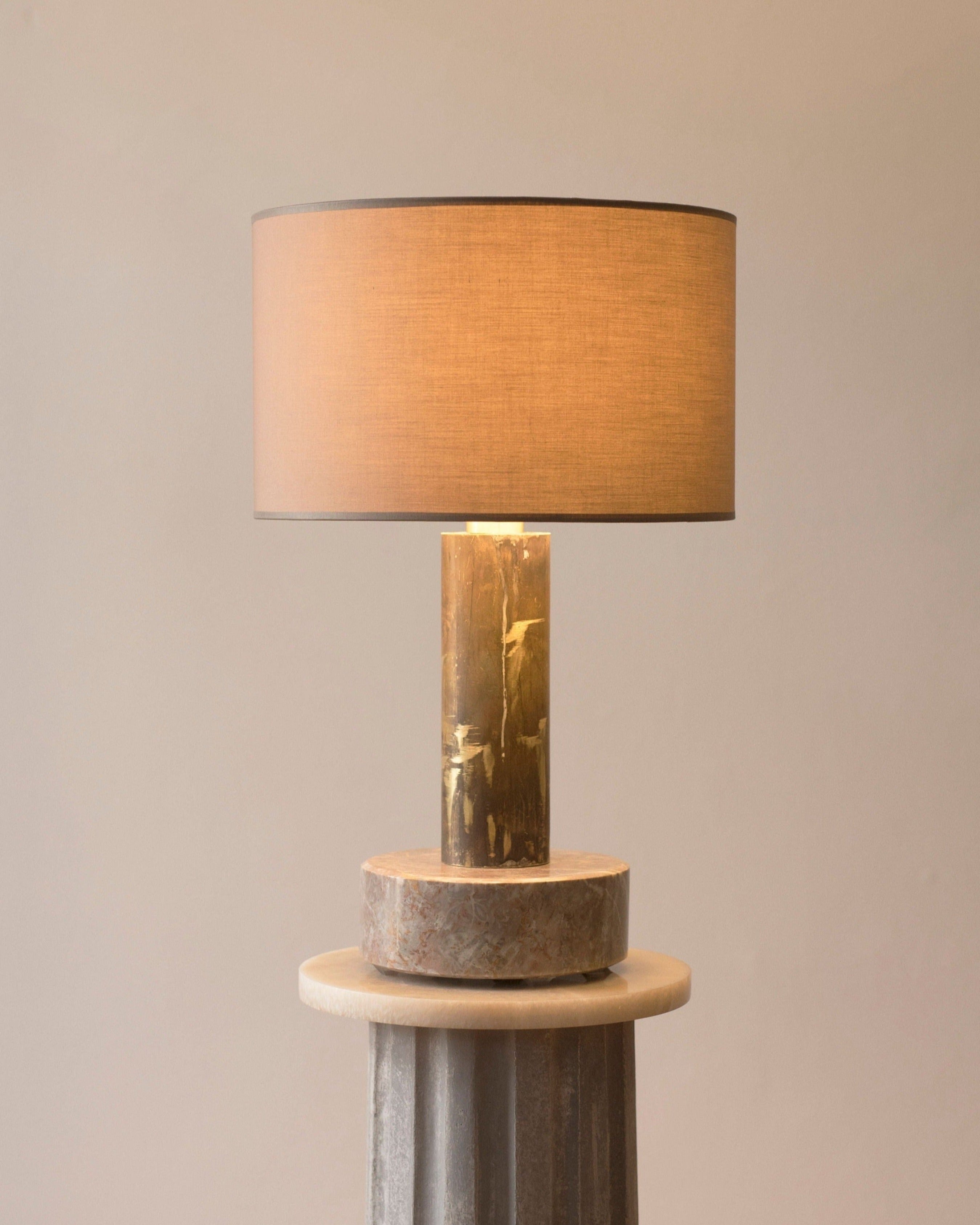 A modern dimmable Brajak Vitberg Pompeii Table Lamp 3 with a cylindrical fabric shade and a polished, marble base, set on a cylindrical pedestal against a neutral-colored wall.