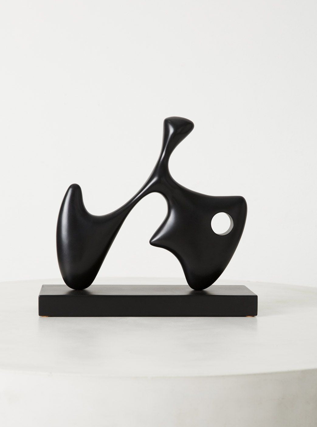 A sleek, black Dela wood sculpture with a smooth surface and an organic shape, featuring a central circular hole, displayed on a grey pedestal against a plain white background by Chandler McLellan.
