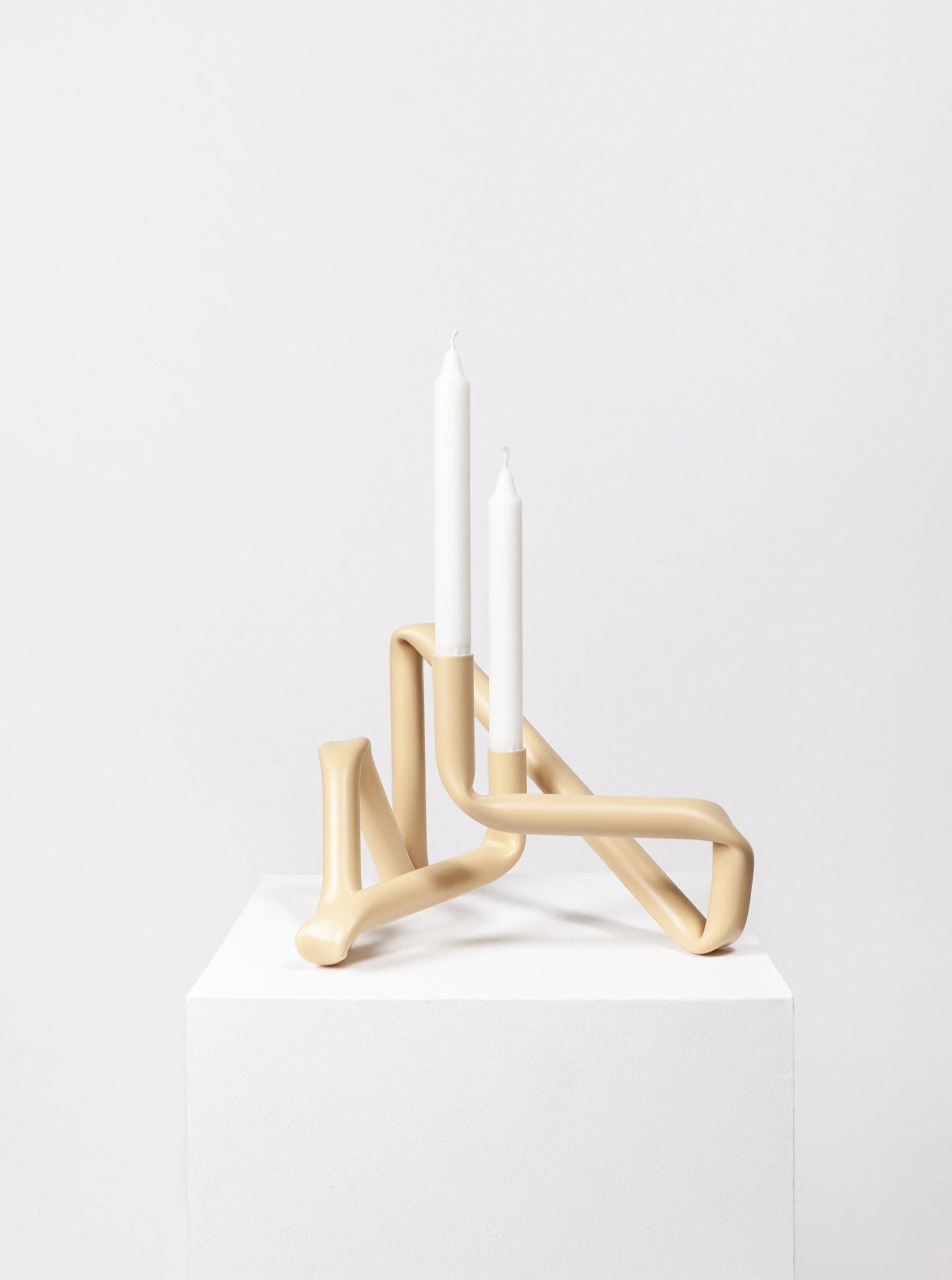 The Bucatini Candle Holder from AOT STUDIO is a modern masterpiece featuring a beige tubular design that holds two white taper candles. Placed on a white pedestal against a light gray background, this candle holder highlights Danish craftsmanship with its unique, twisted handmade design.