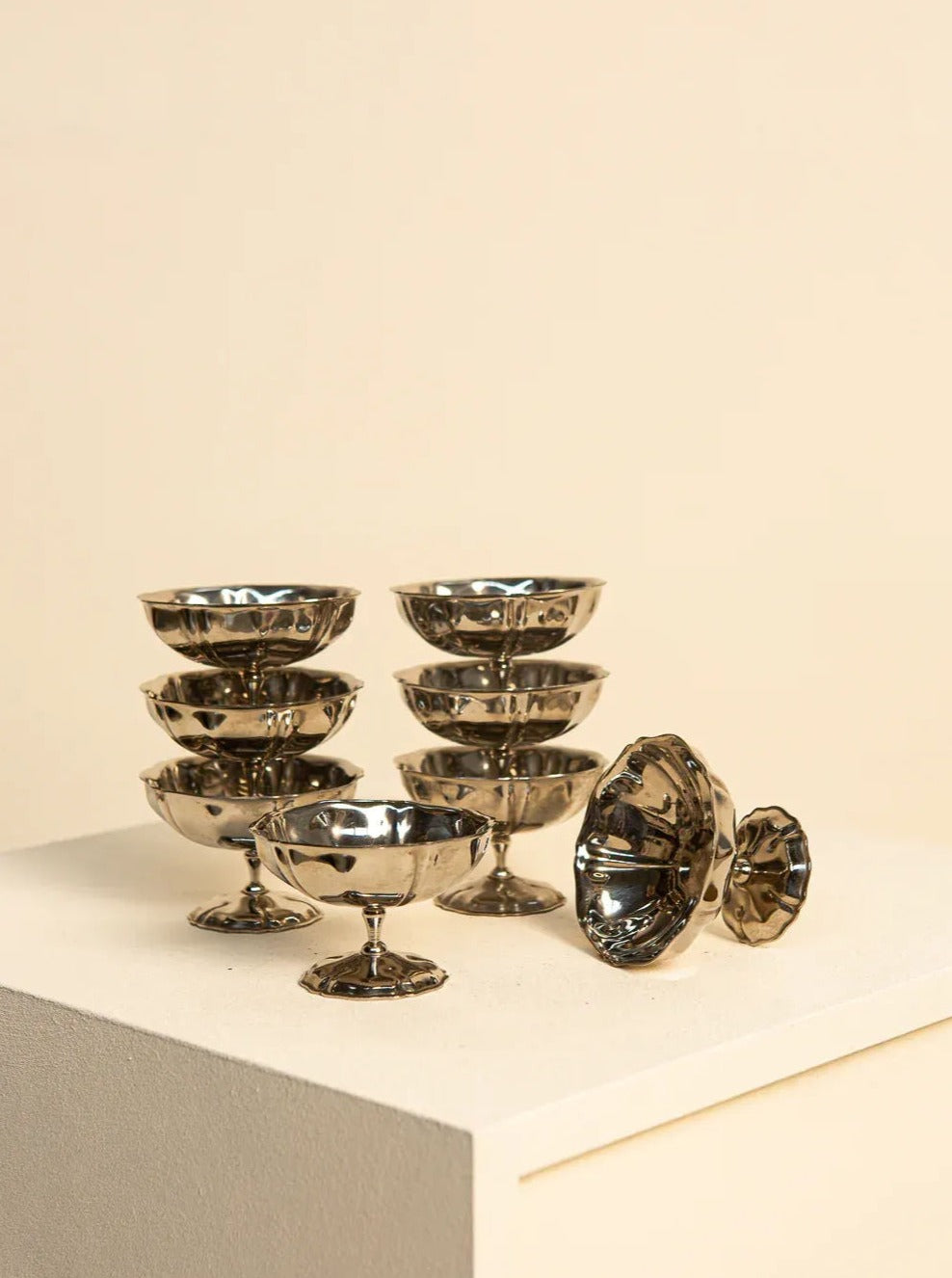 A Treaptyque Set of 8 - 70's Stainless Steel Ice Cream Bowls with a retro design are stacked in two neat piles on a white pedestal. Two additional bowls, showcasing French artisanal craftsmanship, are placed beside the stacks—one upright and the other tipped over, revealing their reflective interiors. The background is a plain beige wall.