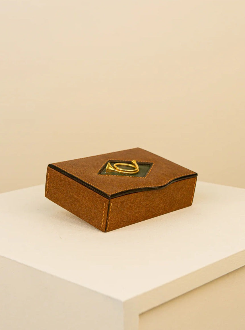A brown Treaptyque leather box with a golden clasp, showcasing a metallic letter "a" on top, placed on a white pedestal against a beige background.