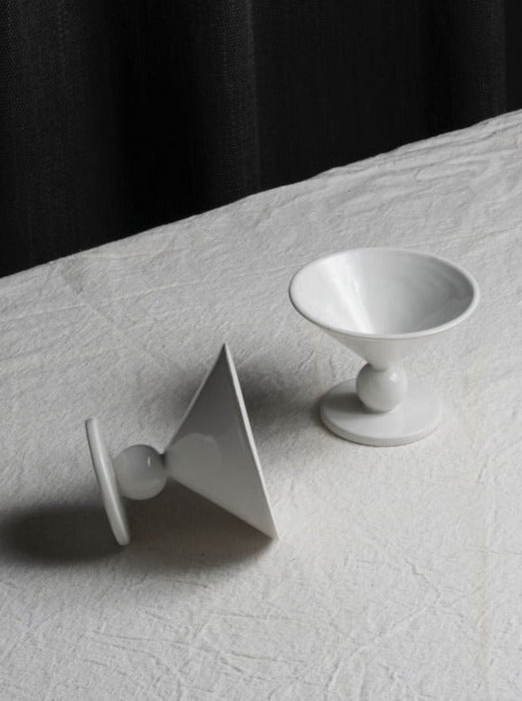 A set of two Les Objets Amsterdam Memphis Ice Cups placed on a light-colored, textured fabric. One cup stands upright, while the other is lying on its side. The backdrop features a dark curtain casting shadows across the fabric. Ideal for those who appreciate the charm of vintage ceramic goblets with a classic touch.