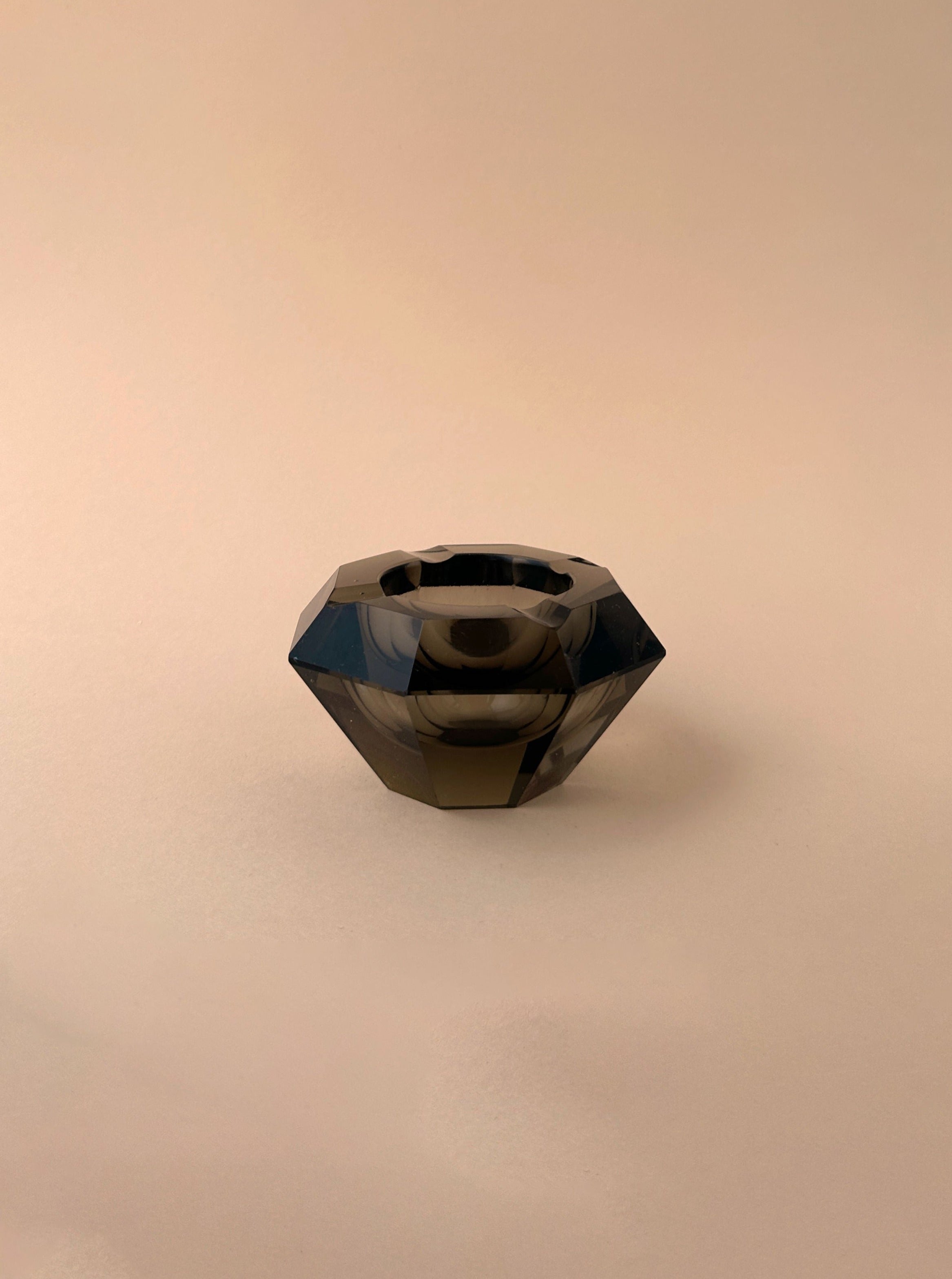 The Moser Crystal Glass Ashtray Prisma 30s from Dodo Vintage is a faceted, dark, crystal-like object with a circular, hollow center. It has a shiny surface with reflections and stands on a plain, beige background. The prism-cut design gives it an elegant resemblance to both an Art Deco ashtray and a decorative gemstone-like item.