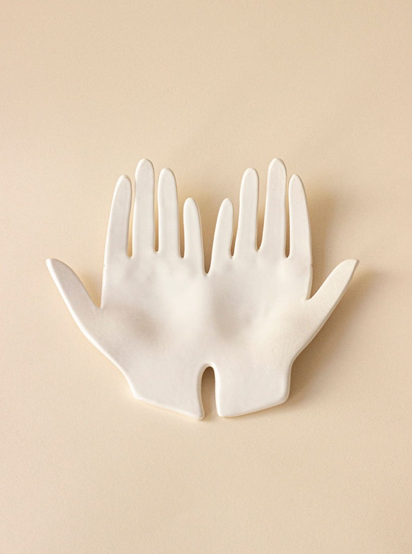 A Main Edition ceramic wall art piece depicting two overlapping hands, with a smooth, matte finish, mounted on a delicate sandstone background.