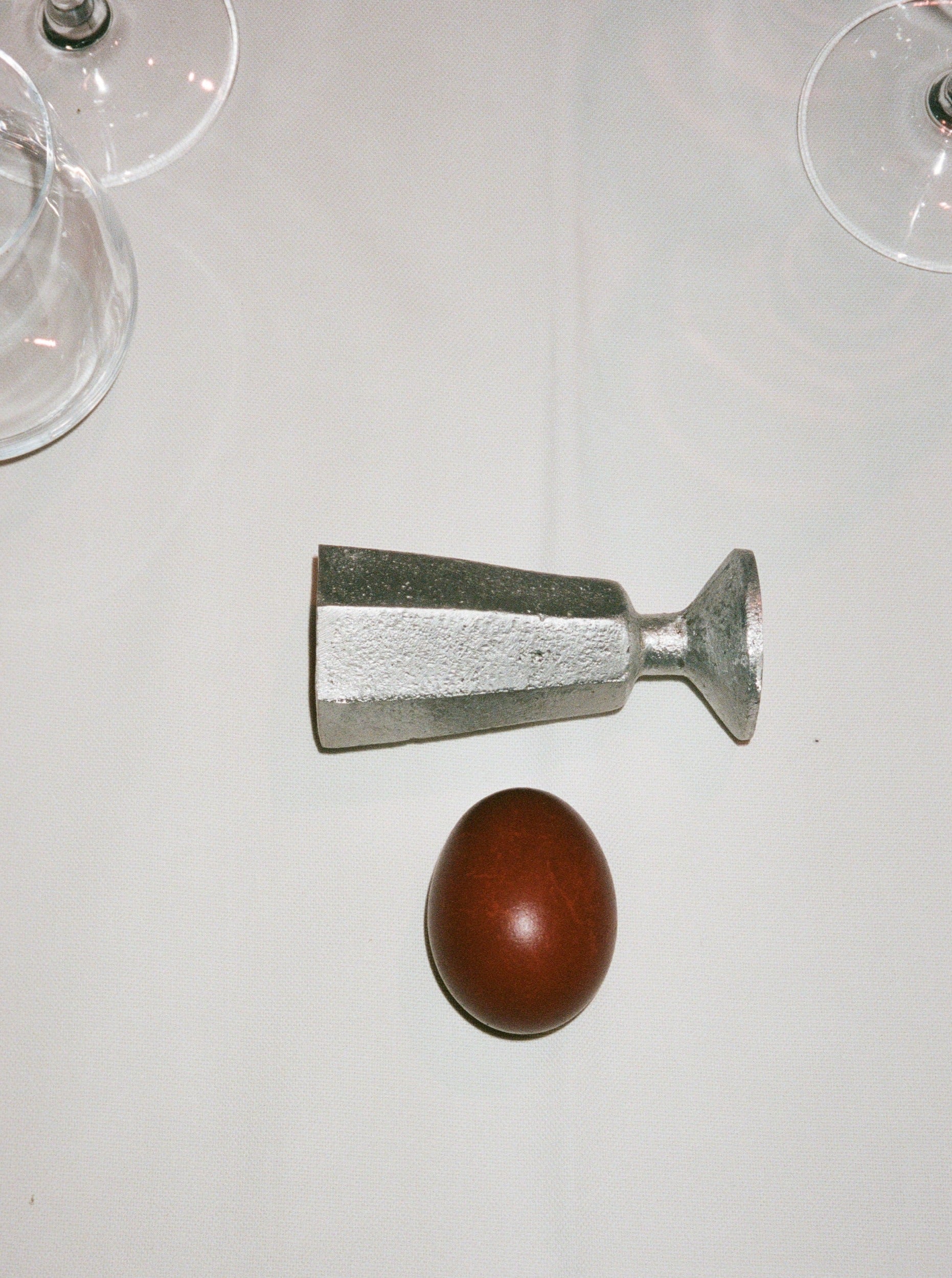 A Rooms Studio high-grade aluminum egg holder lying next to a brown egg on a white tablecloth, with two glass stems partially visible at the edges.
