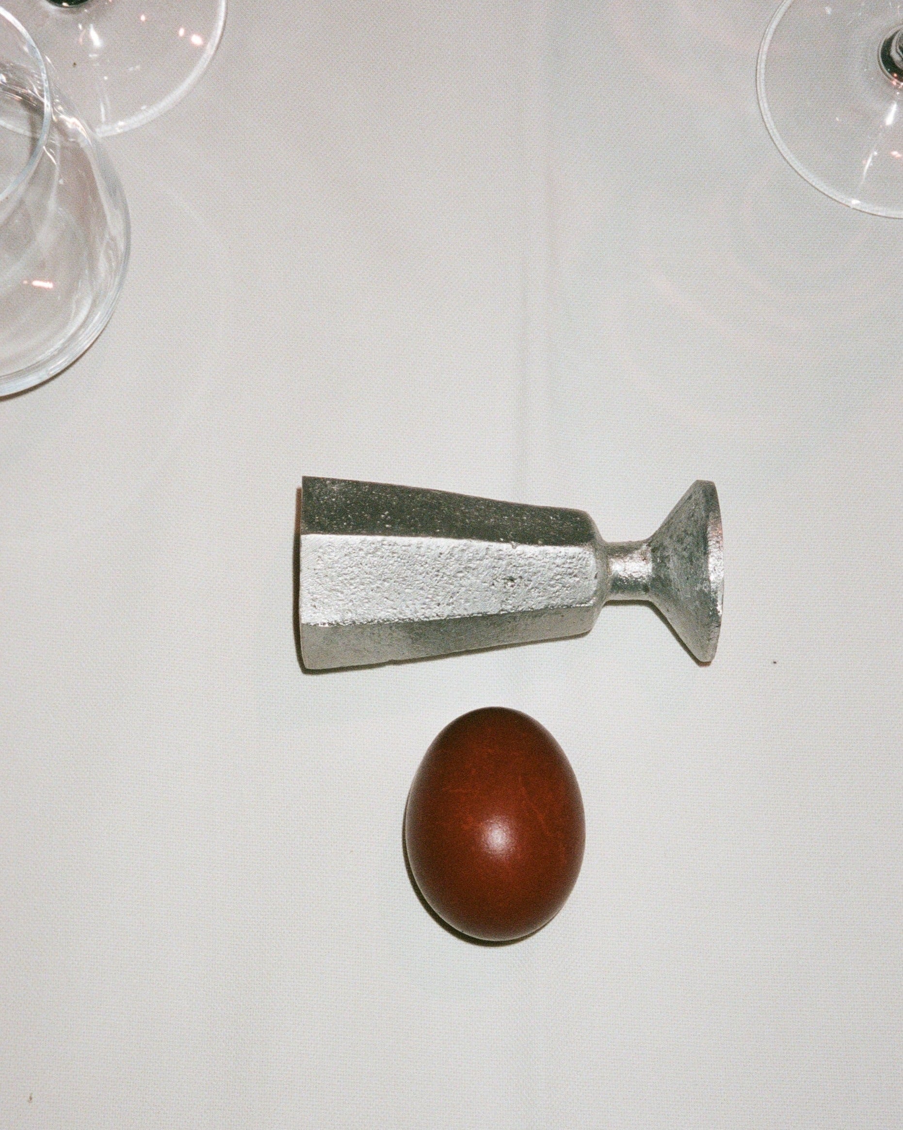 A Rooms Studio high-grade aluminum egg holder lying next to a brown egg on a white tablecloth, with two glass stems partially visible at the edges.