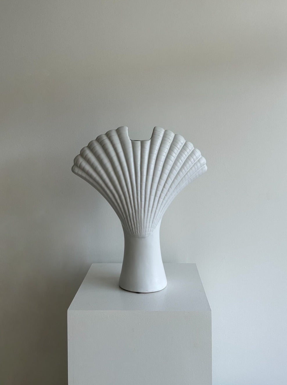 A white, shell-shaped ceramic vase from Les Objets Amsterdam, known as the Shell Vase, with a textured surface sits atop a plain white pedestal against a light gray background. The vase's unique design, reminiscent of Vintage Hollywood Regency style, resembles a fan or scallop shell with a narrow base flaring outwards.