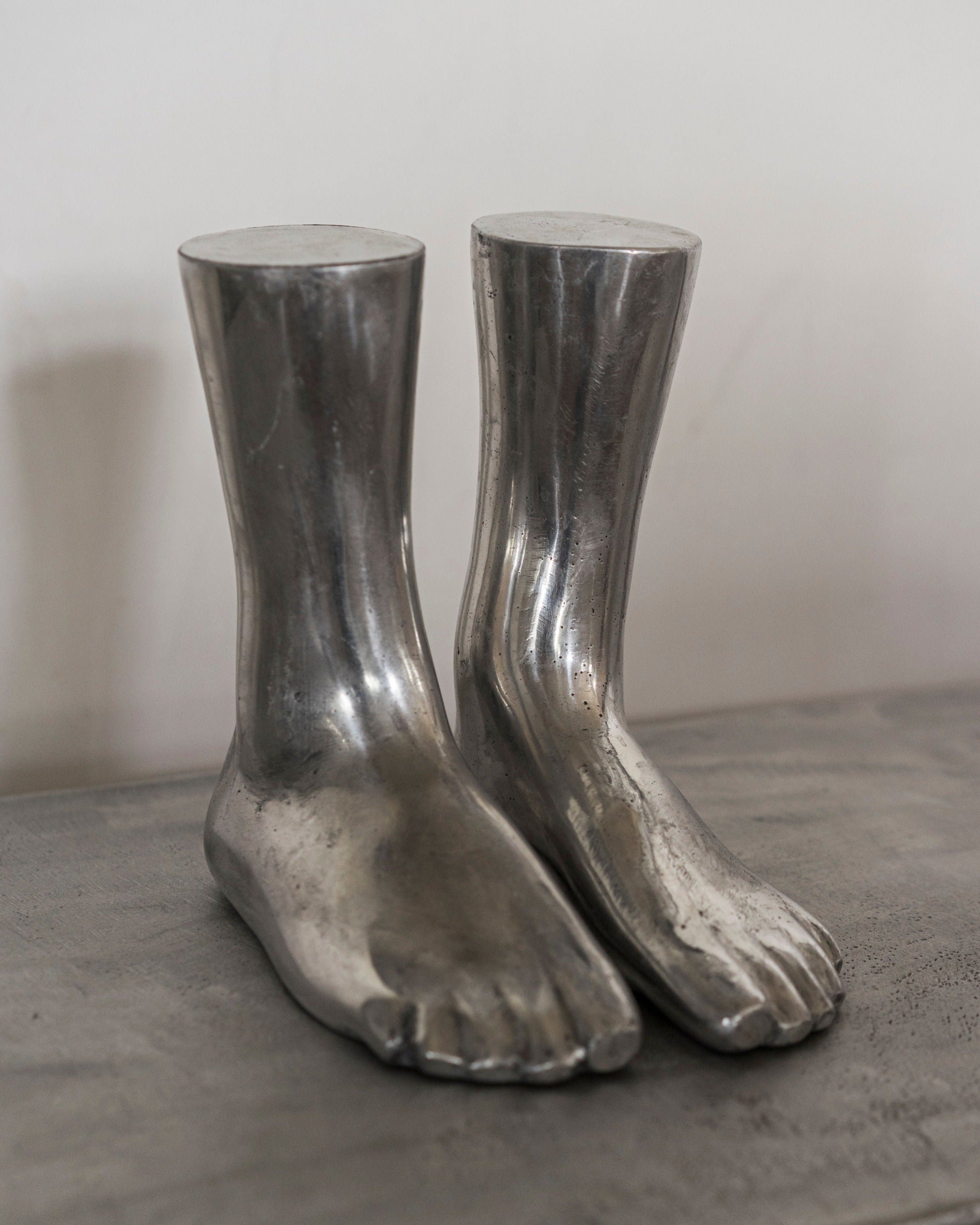 A pair of Rooms Studio casted aluminum feet sculpture boots designed to resemble human feet, with detailed toes and shin-high length, displayed on a grey surface against a neutral background.