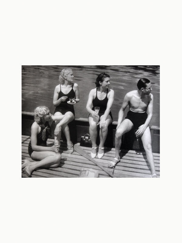 Black and white photo of four people in vintage swimwear sitting by a wooden dock; three women and one man enjoying a sunny day, one woman holding a drink, reminiscent of the Maison Plage art scene with Brassai: Paris & Picasso.