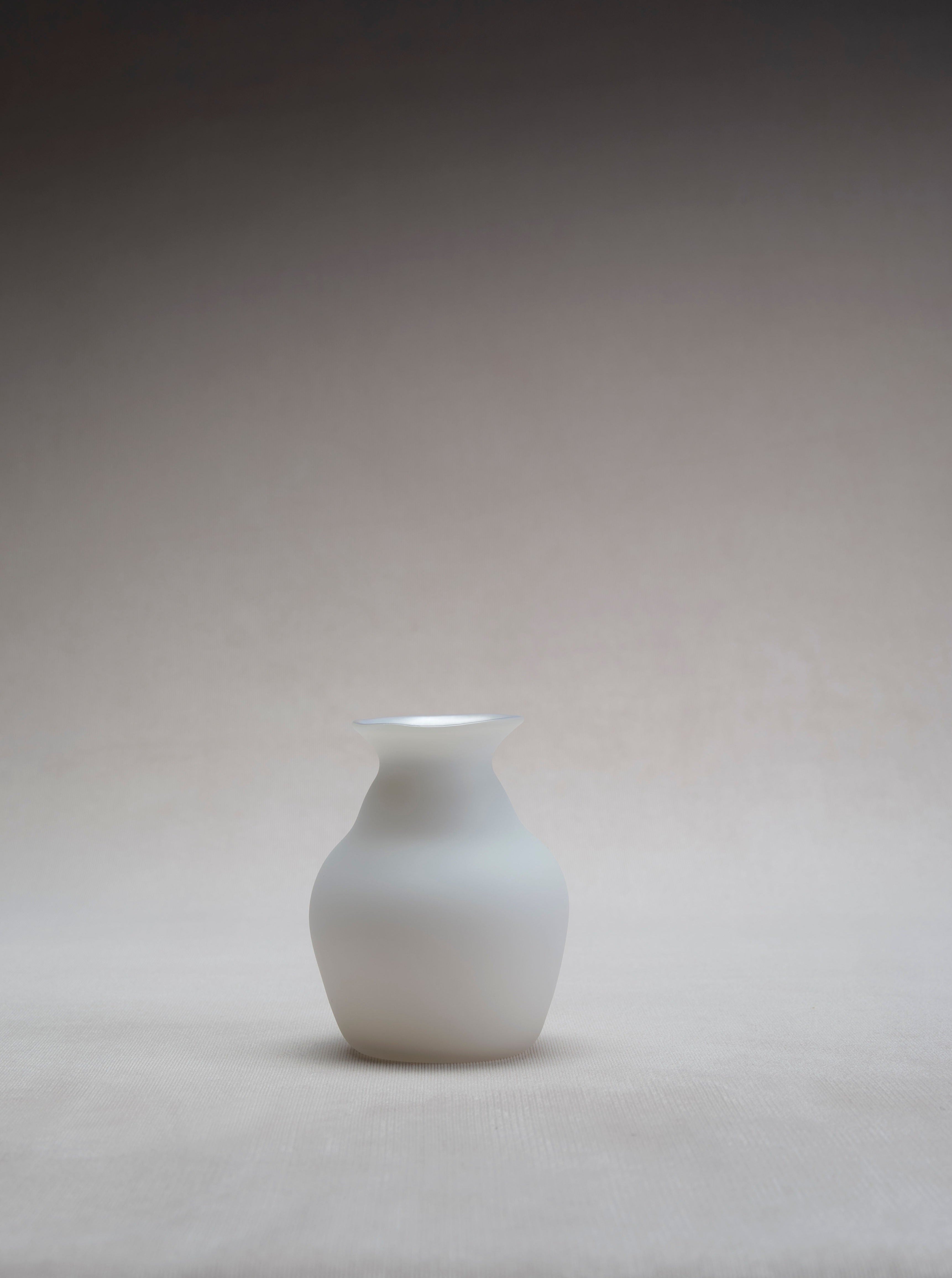 A simple, white ceramic **Small Vase in Cotton** by **Verre d'Onge** is placed against a neutral, gradient background that transitions from light beige at the bottom to a darker tone at the top. The small vase has a smooth, matte finish and a classic, rounded shape with a narrow neck, making it an elegant piece of home decor.