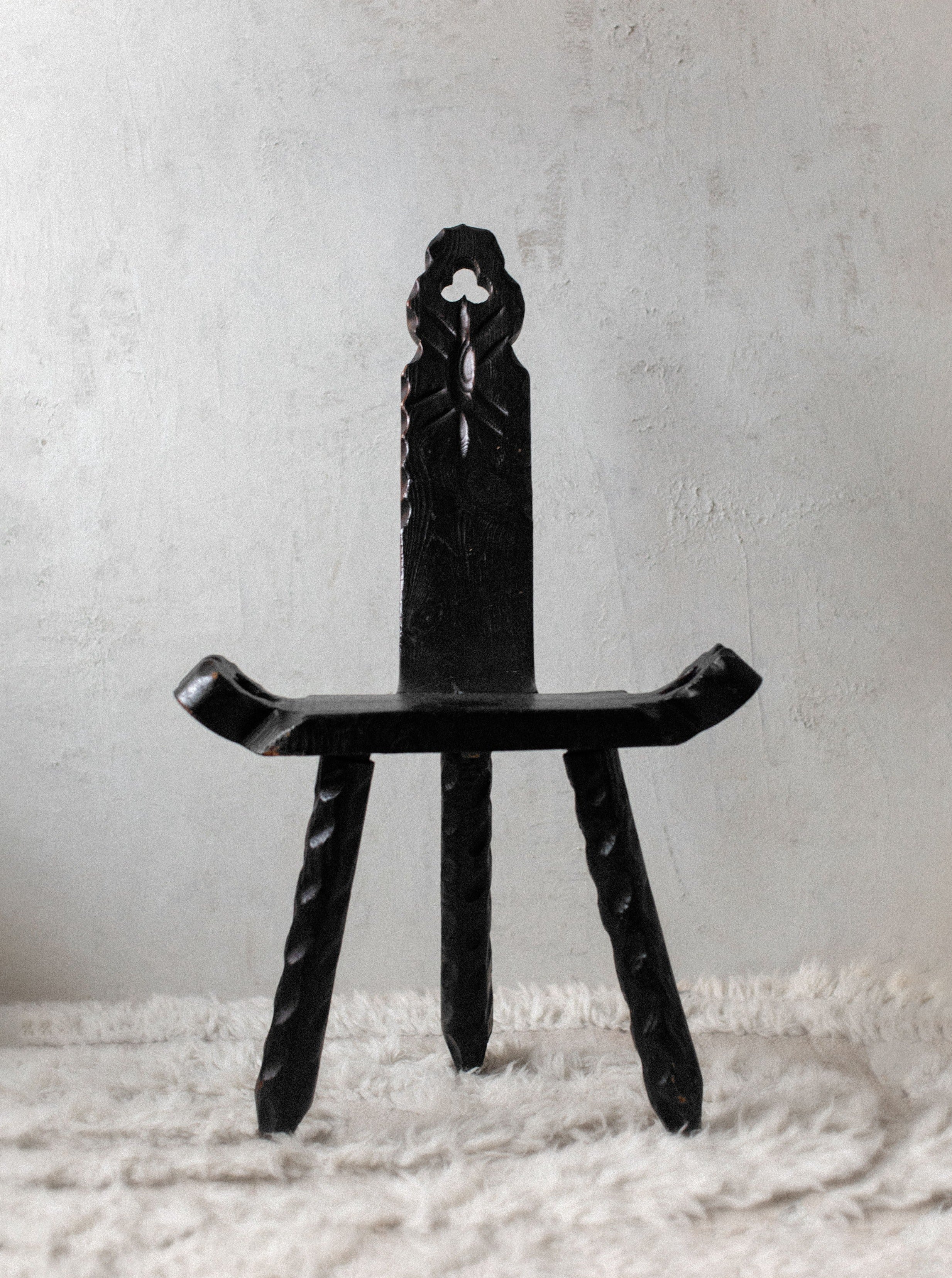 A black, ornate Out For Lunch Brutalist chair with intricate carvings, including a figure at the top of the backrest, stands on a rugged textured rug against a plain white wall.