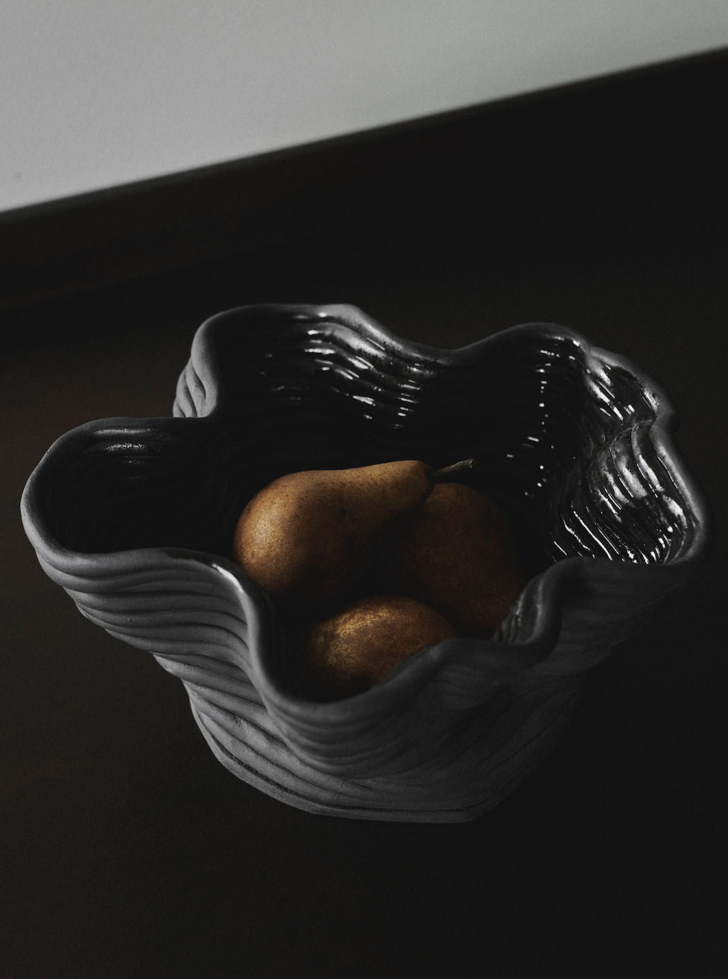 The 2222Studio Wobbly Bowl Black, with its wavy rim, cradles three pears in a contemporary design. In moody lighting, shadows enhance the texture of both the ceramic bowl and the fruit's surface.