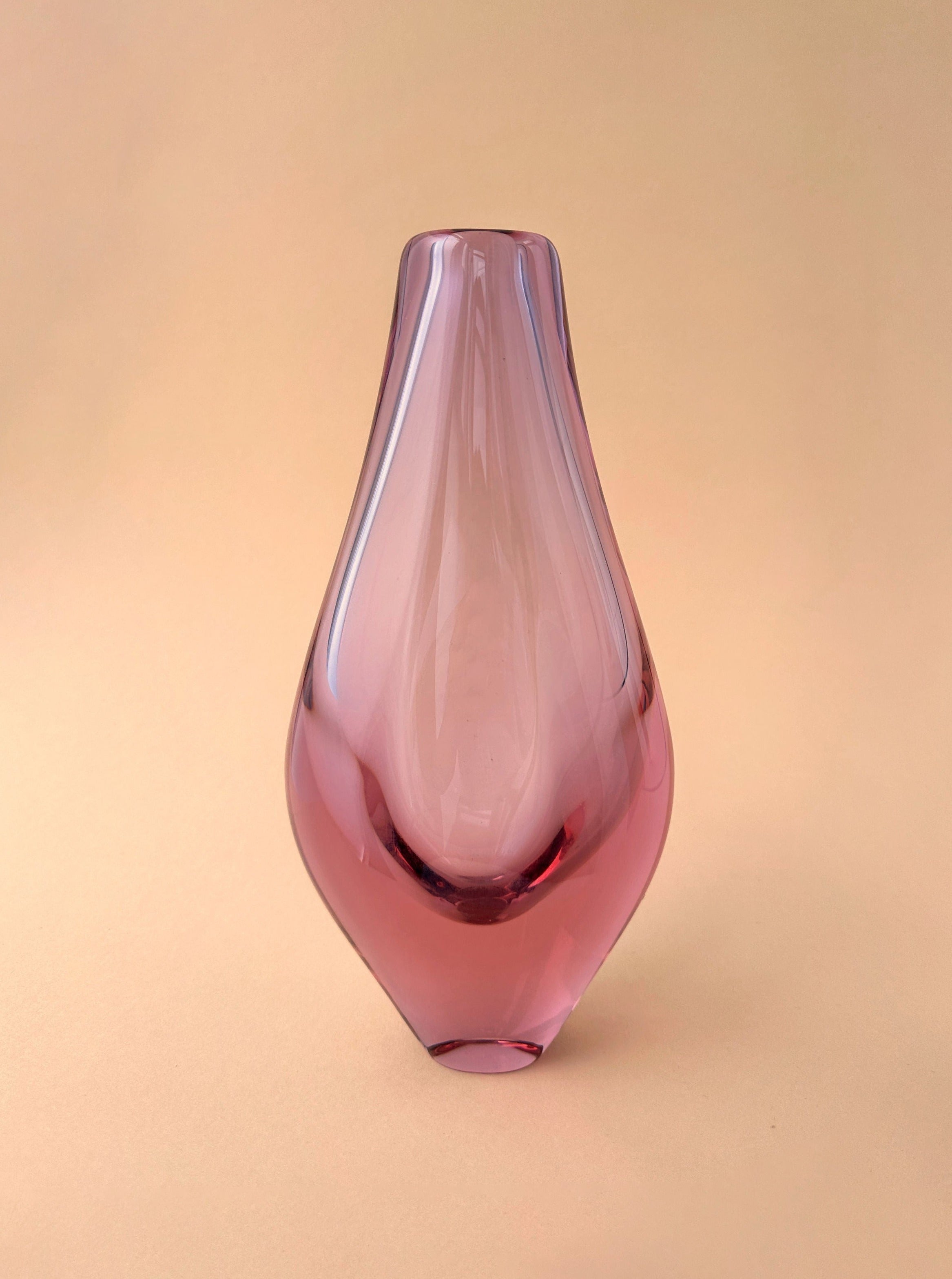 A delicate pink Bohemian Alexandrite glass vase with a sleek, elongated teardrop shape, displayed against a soft peach background.