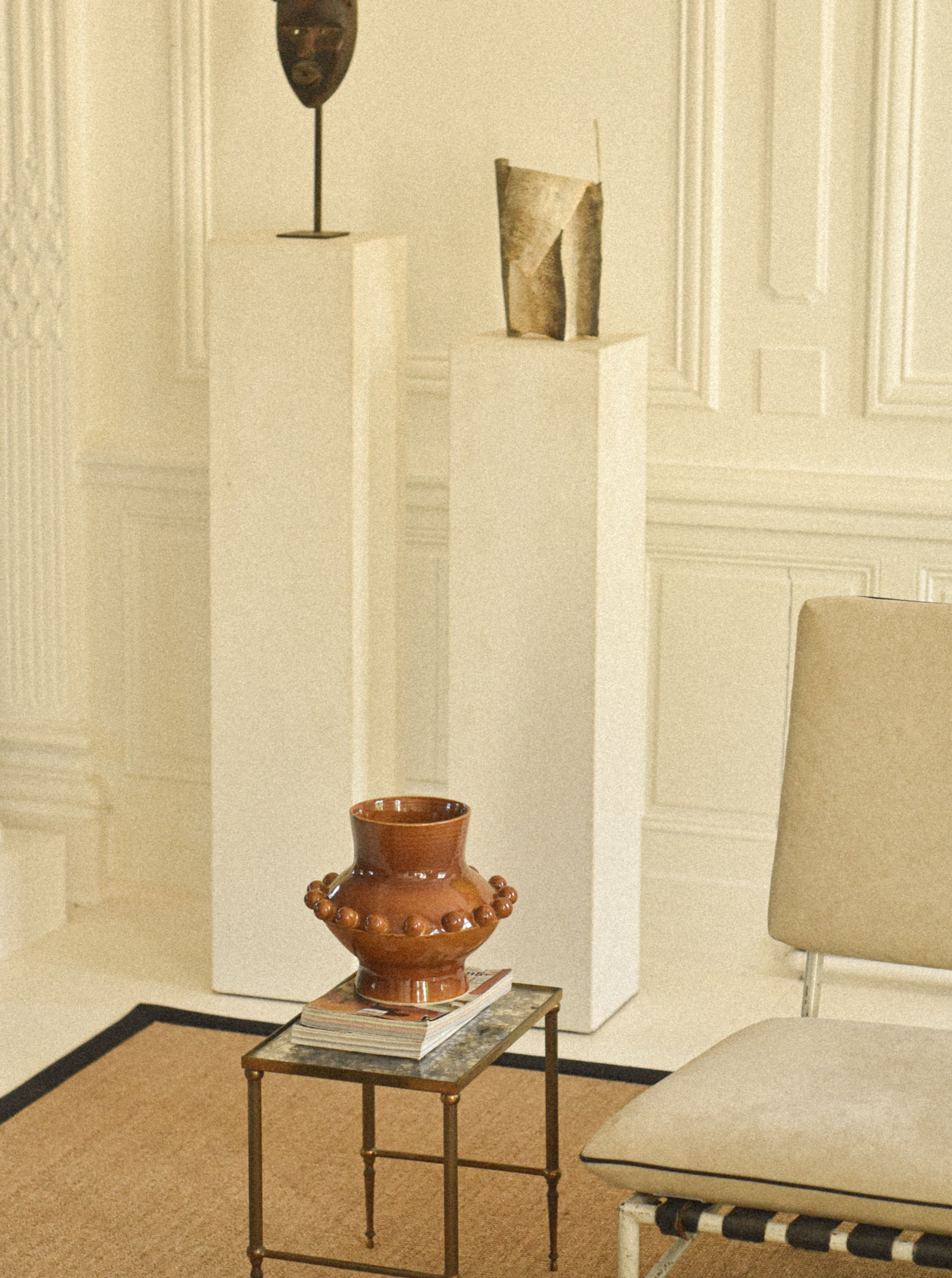 An elegant interior featuring three tall white pedestals displaying abstract sculptures, with a unique Landy Rakoto Ceramics UFO Vase in Amber placed on a small glass table. The room has a classical design with detailed wall molding.