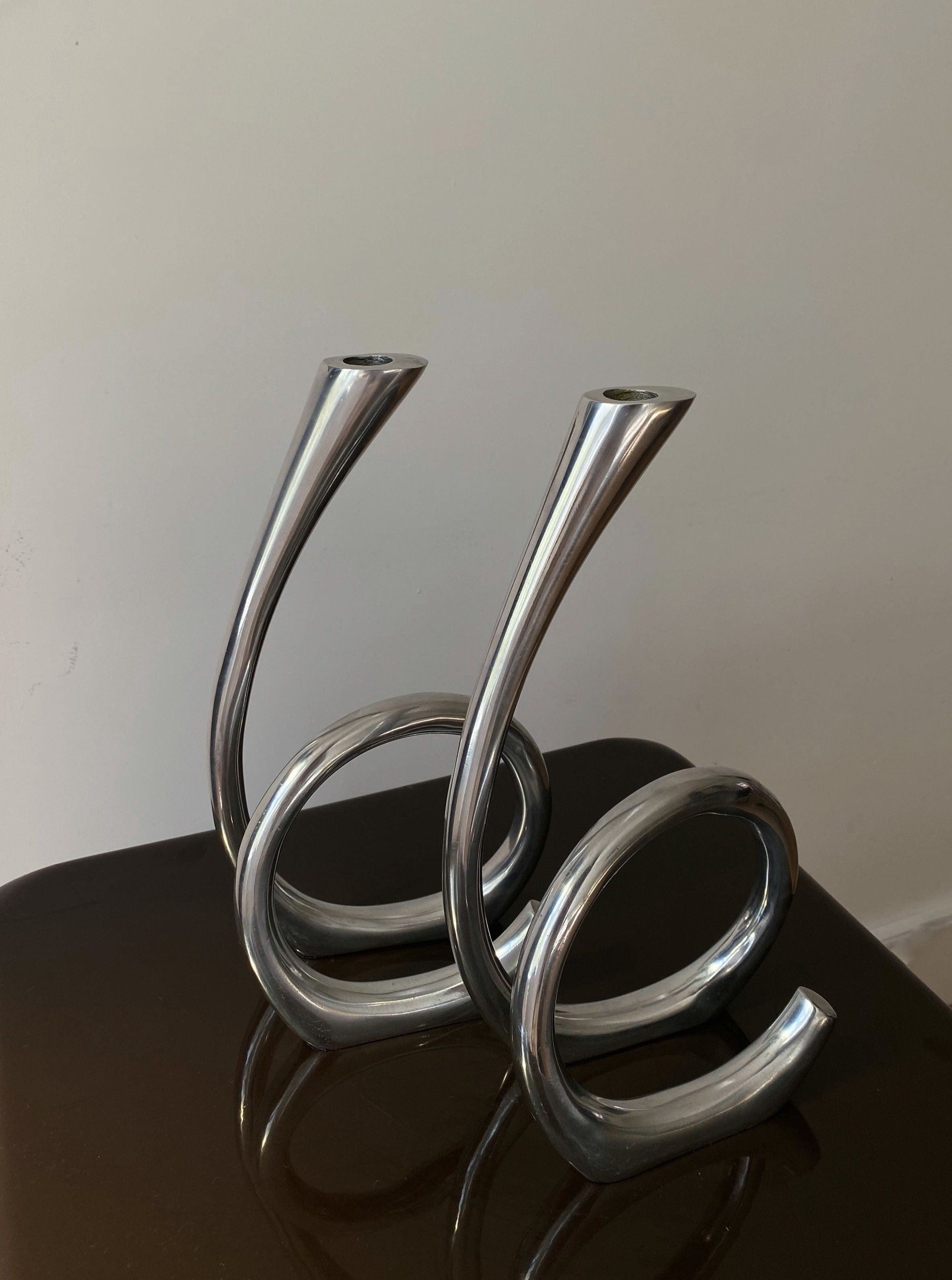 Two Vintage Curly RVS Candle Holders from Les Objets Amsterdam with a sleek design standing on a brown surface against a light-colored background.