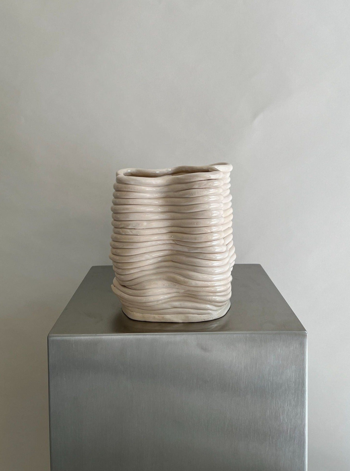 A textured handcoiled ceramic Wobbly Vase Natural from 2222Studio with a unique, wavy design sits atop a gray metal pedestal against a neutral gray background.