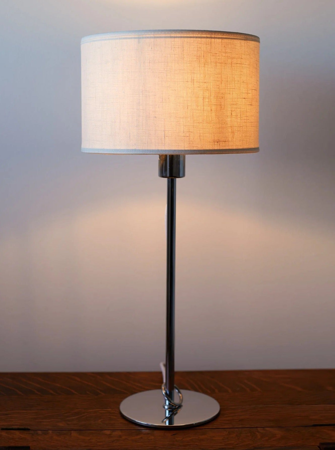 A contemporary Chrome Table Lamp with a cylindrical beige shade, standing on a chrome base placed on a wooden surface, lit and casting a warm glow against a neutral wall by Out For Lunch.