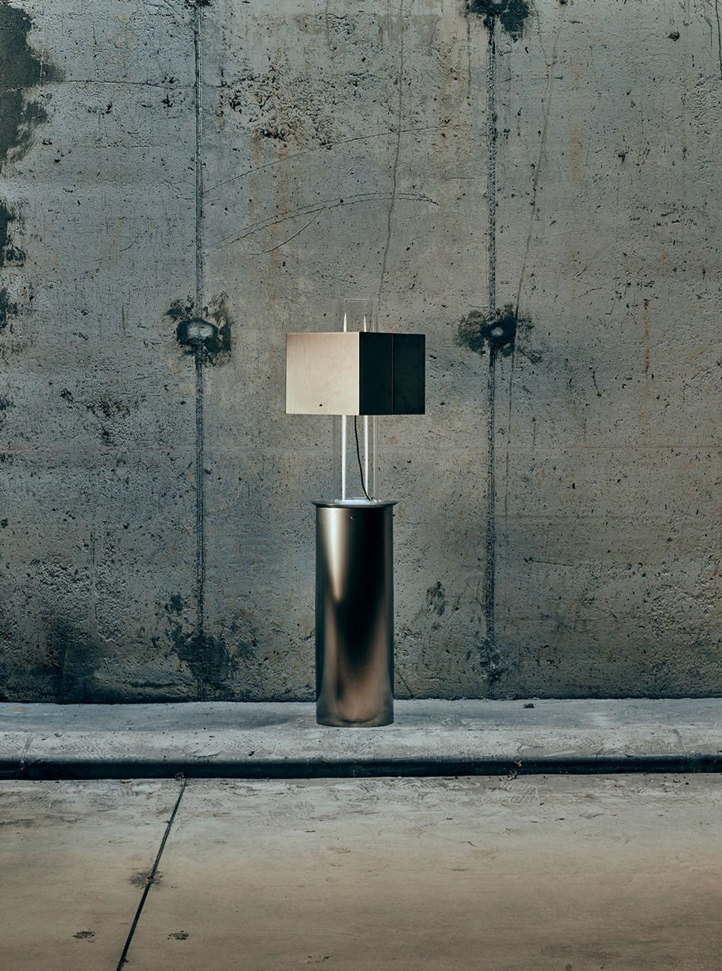 The Metal Floating Table Lamp by Brajak Vitberg features a geometric design on a cylindrical base against a textured concrete wall, enhancing its industrial and minimalist aesthetic for a modern feel.