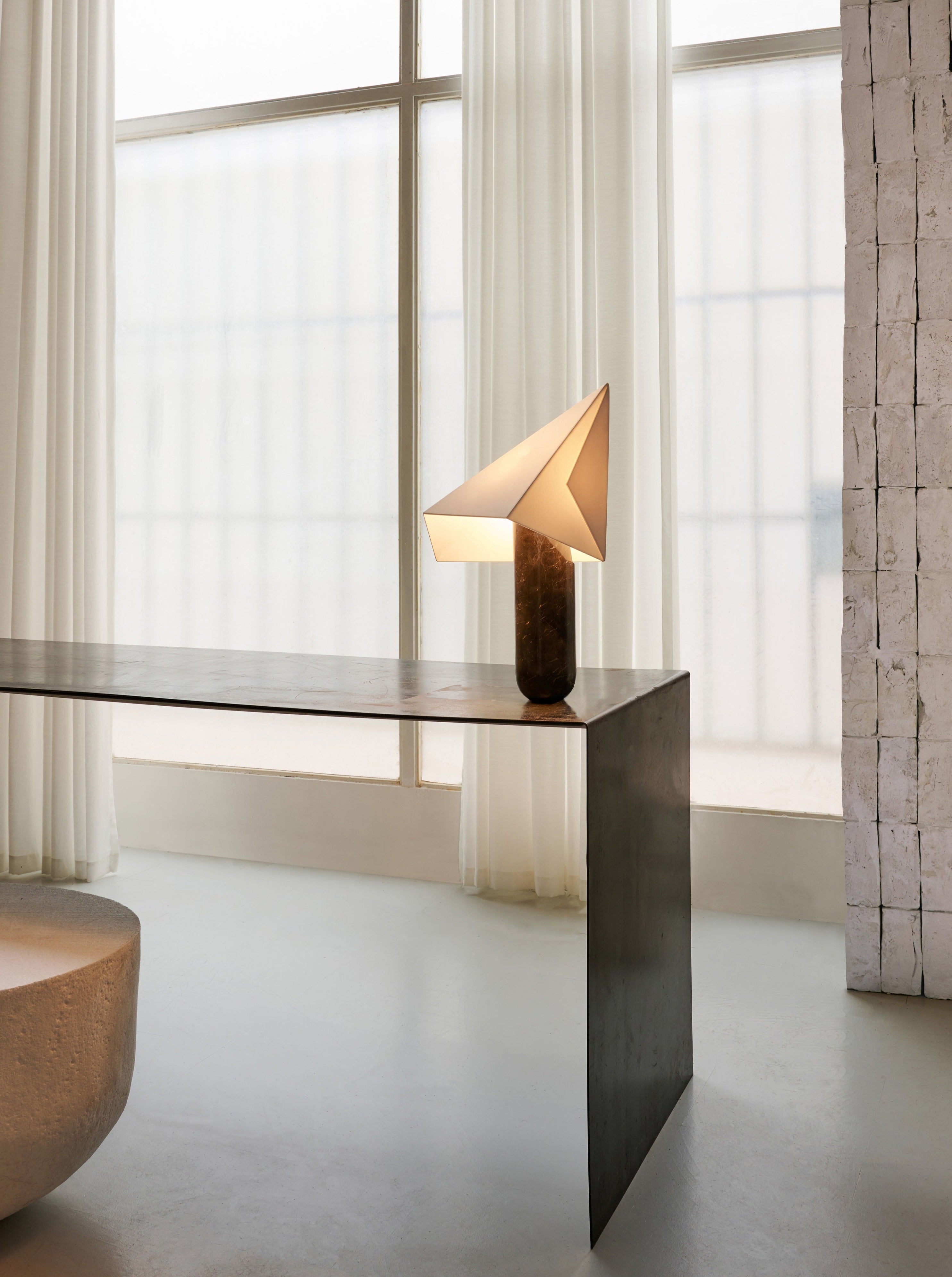 A minimalist interior with a sleek metallic table, featuring an Acme - Greek Marble Black table lamp on top, near sheer white curtains and large windows allowing natural light, designed by Mariza Galani.