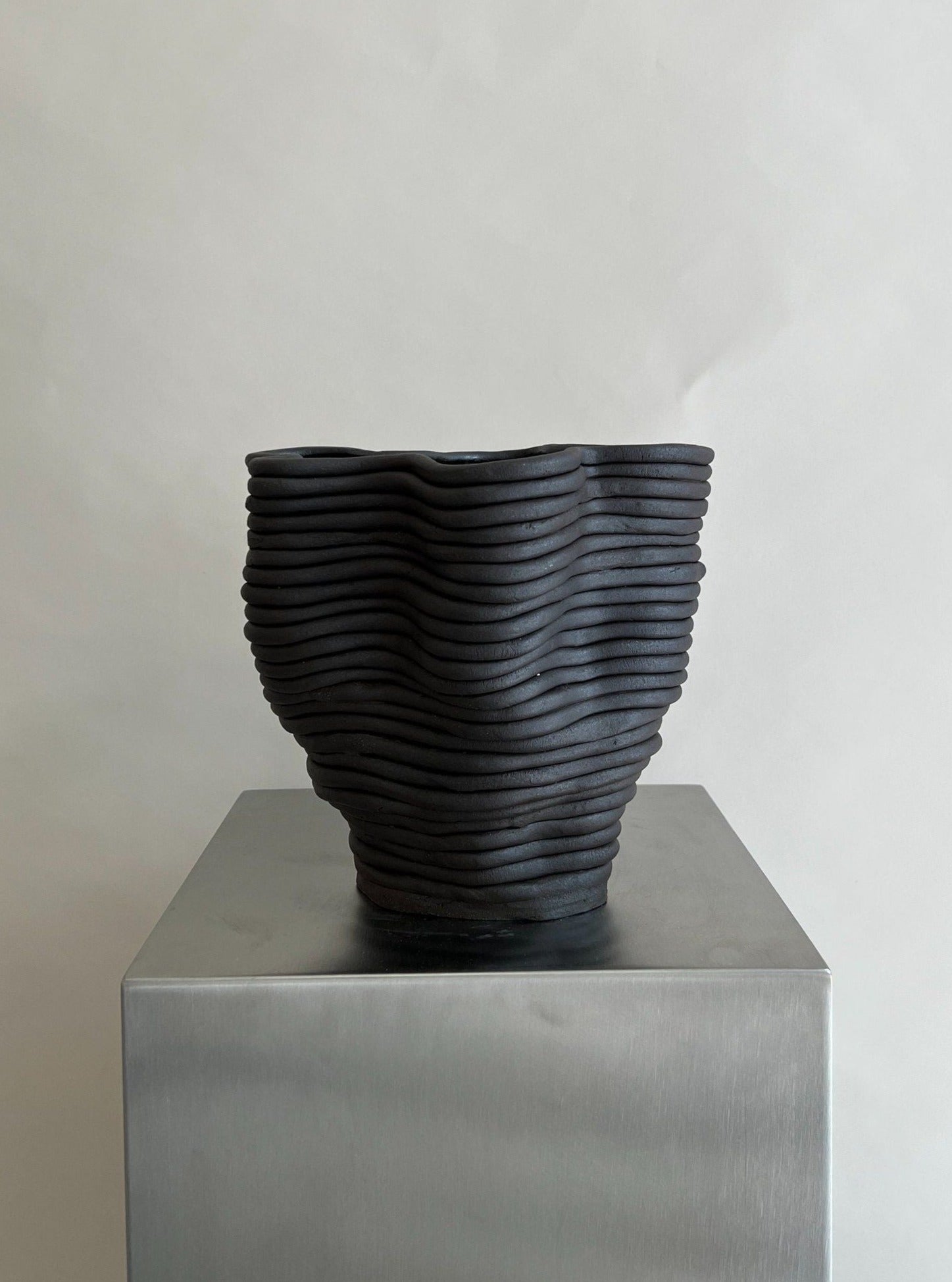 The Wobbly Vase Black by 2222Studio, with its curvy ribbed design and organic flowing shape, rests elegantly on a shiny metallic pedestal against a plain white background, showcasing its sculptural ceramic form and textured layered surface that enhance its minimalist aesthetic.