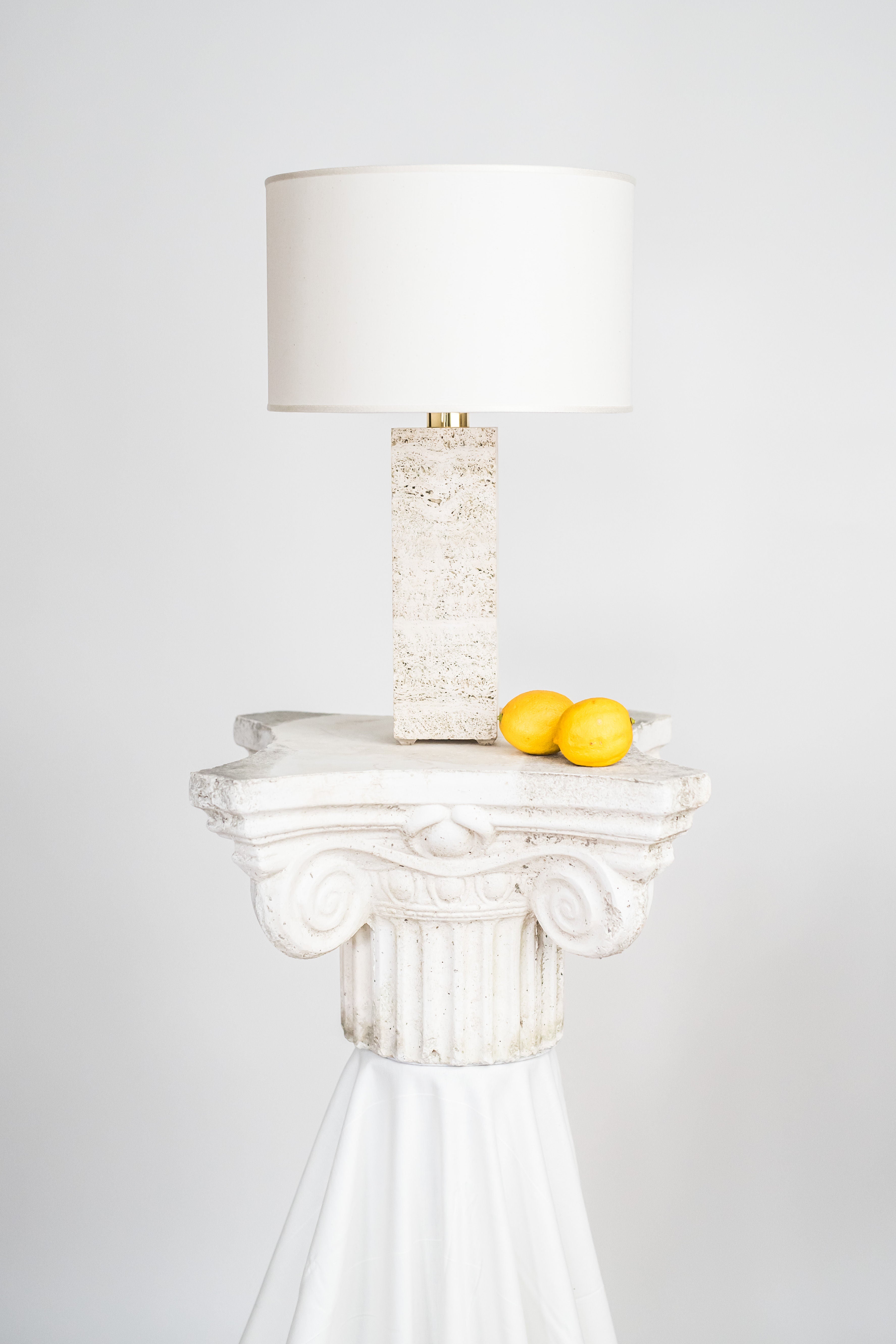 A Capri Travertine table lamp 1 by Brajak Vitberg with a white shade and a unique travertine body sits on an ornate white classical column pedestal, which holds two yellow lemons on a draped white cloth. The background is plain.