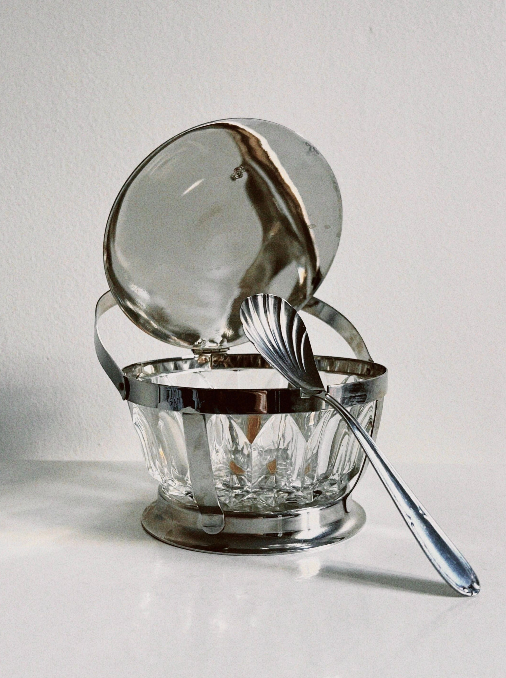 A vintage Manushi Crystal Sugar Bowl with a hinged lid, made of clear cut glass and trimmed in silver, sits on a white surface. The lid is open, and a silver spoon with an ornate handle rests inside the bowl. The background is a plain, off-white wall.