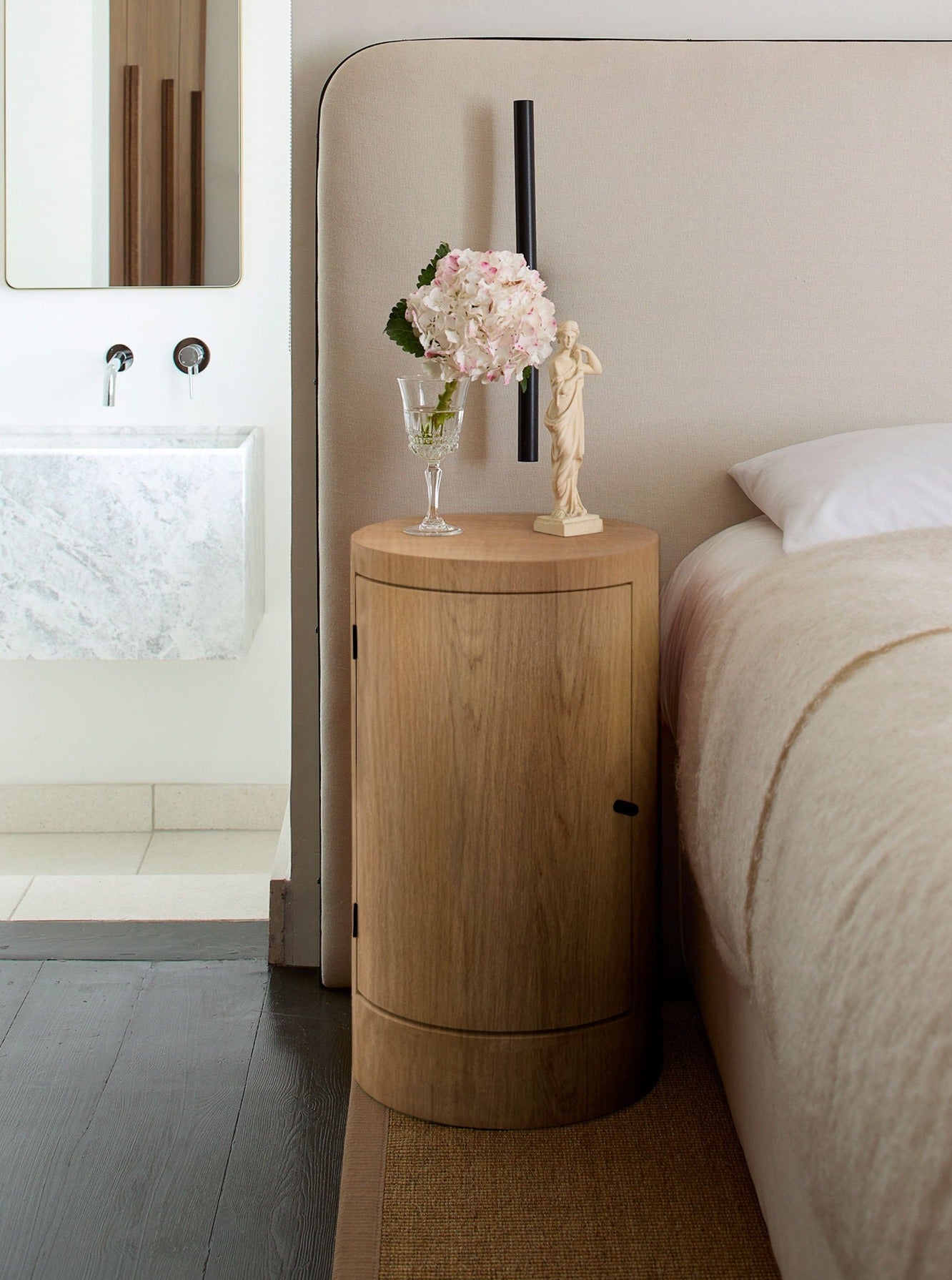 A modern bedroom features a pair of Constant Nightstands in Oiled Oak by Lemon beside a beige upholstered bed. The nightstands hold a clear vase with pink flowers and a small, elegant statue. A minimalist bathroom with a marble sink is partially visible in the background.