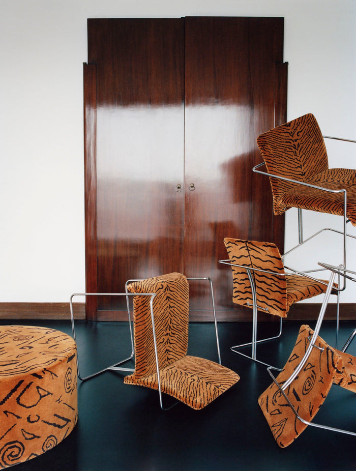 A playful arrangement of chairs and furniture featuring bold tiger and tribal patterns on upholstery, juxtaposed against sleek chrome frames and a dark wood-paneled backdrop.