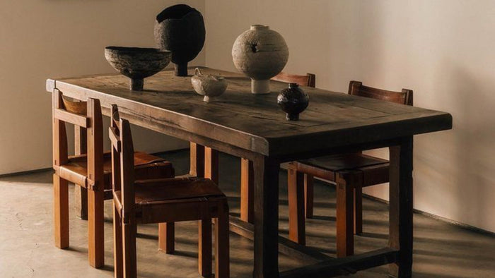 The setting features a robust wooden table with natural imperfections, paired with solid wooden chairs. The muted, earthy tones and minimal decor create a serene yet warm atmosphere, blending contemporary aesthetics with rustic charm.