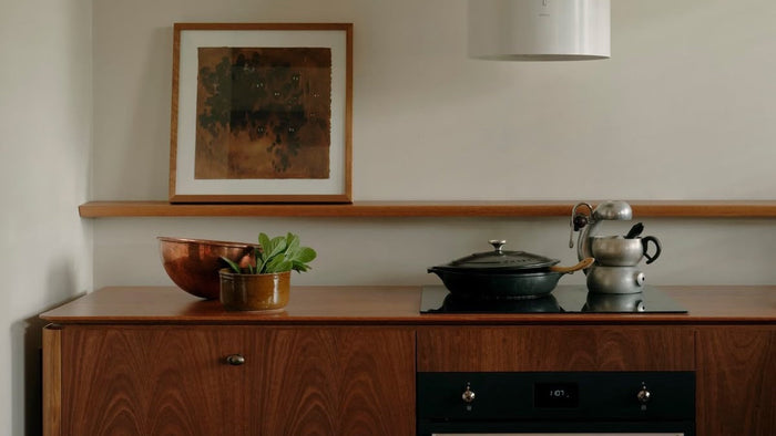 Minimalist kitchen with mid-century modern wooden cabinetry, copper and cast iron cookware, and a framed abstract artwork on a floating shelf. Earthy tones and warm textures create a refined, timeless aesthetic.