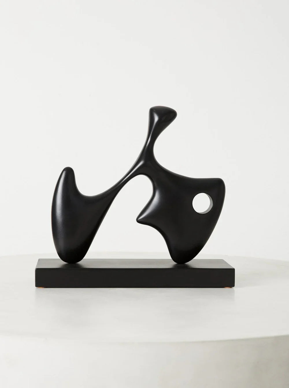 Abstract black sculpture with smooth, flowing shapes and a small circular cutout, mounted on a rectangular base. The artwork features organic, fluid lines and a modern, minimalist design, set against a plain white background.
