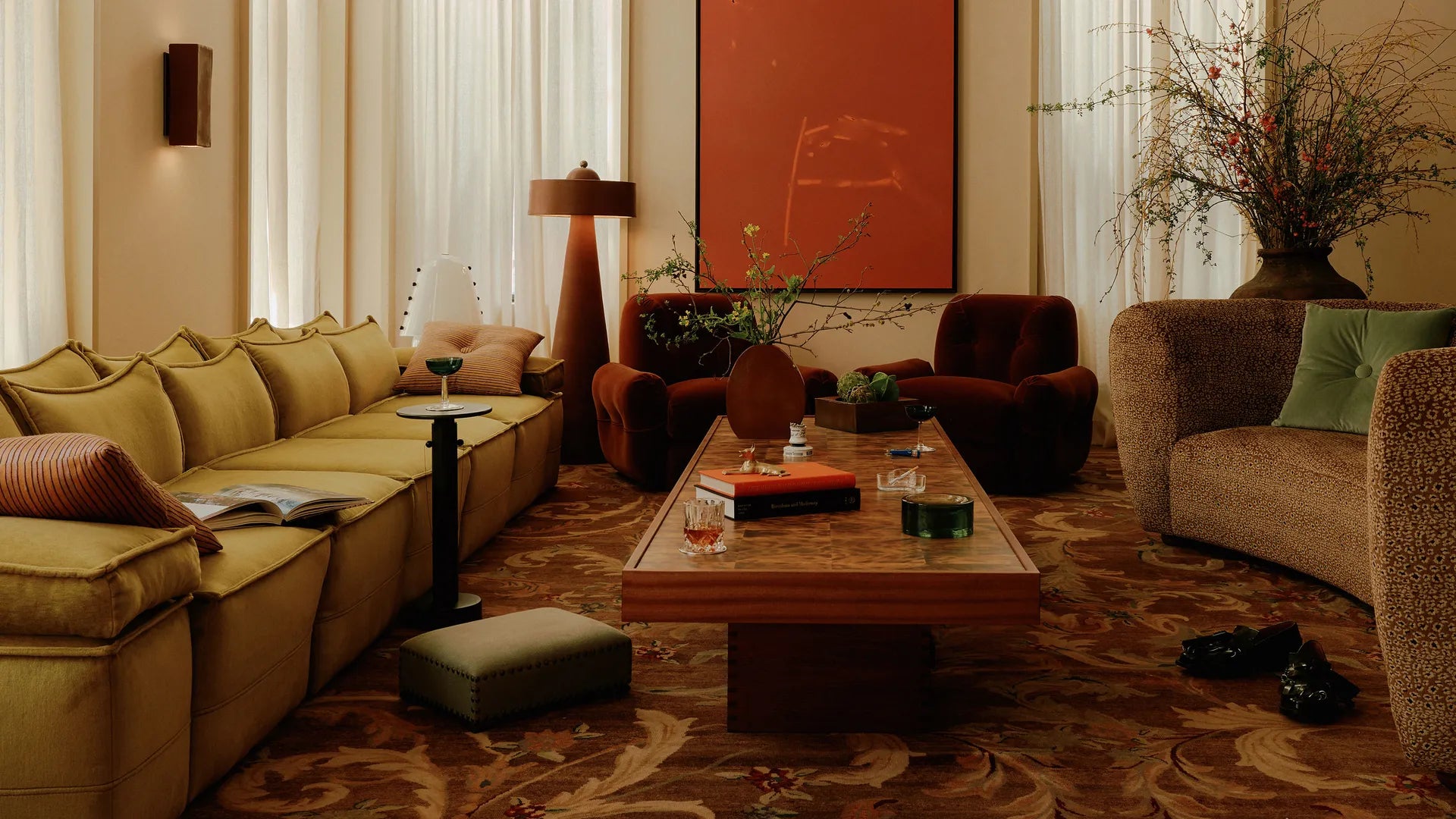 A cozy, stylish living room featuring a mustard-yellow sofa, rust armchairs, and a leopard-print couch. A wood veneer coffee table displays books and decor, while earthy tones, sheer curtains, and bold orange artwork add warmth.