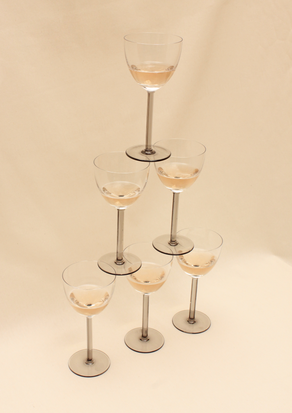 A pyramid of six champagne glasses, with three glasses at the base, two in the middle, and one on top, each partially filled with champagne against a cream-colored background.